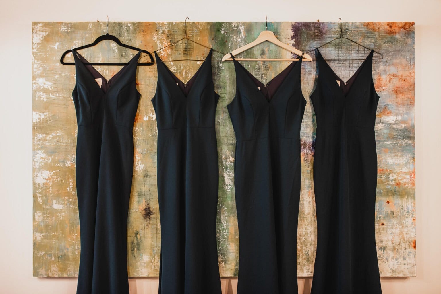 Four black dresses on hangers against an abstract textured painted backdrop, perfect for bridesmaids getting ready at a nearby salon during a Castaway wedding.