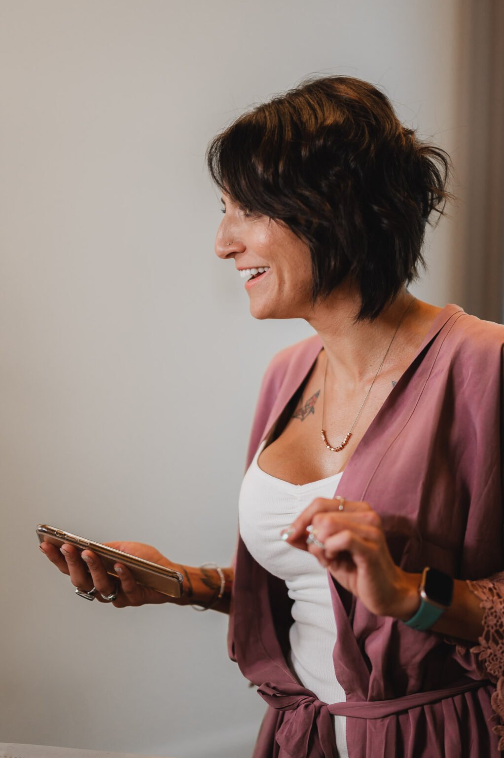 A bridesmaid in a pink cardigan smiles while holding a phone, looking to the left, getting ready at a nearby salon during a Castaway wedding.