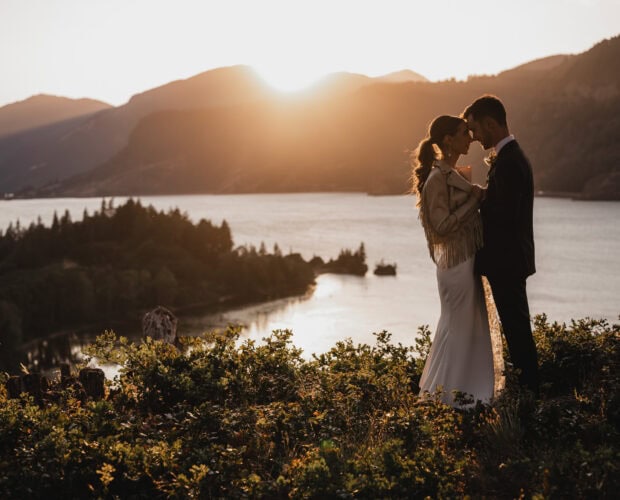 A couple embracing at sunset by a lake, surrounded by hills and foliage against a golden sky, creates a perfect setting for wedding photos at The Griffin House.