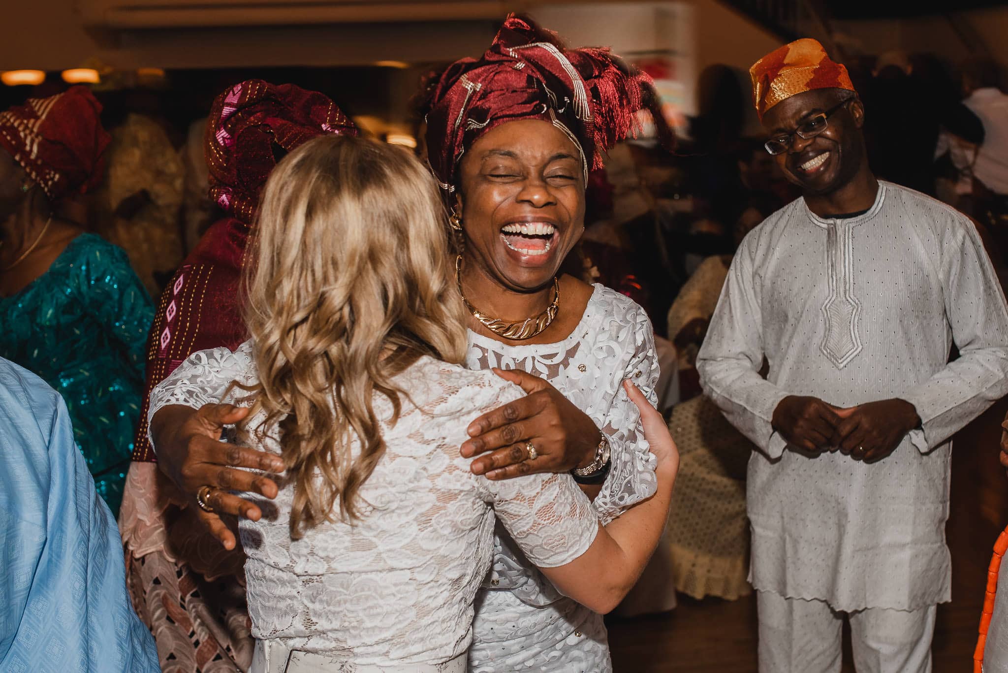 Two women in traditional attire hug and smile joyfully at a vibrant celebration, set against the lively backdrop of a wedding at the Evergreen, with others joining in the festivities.