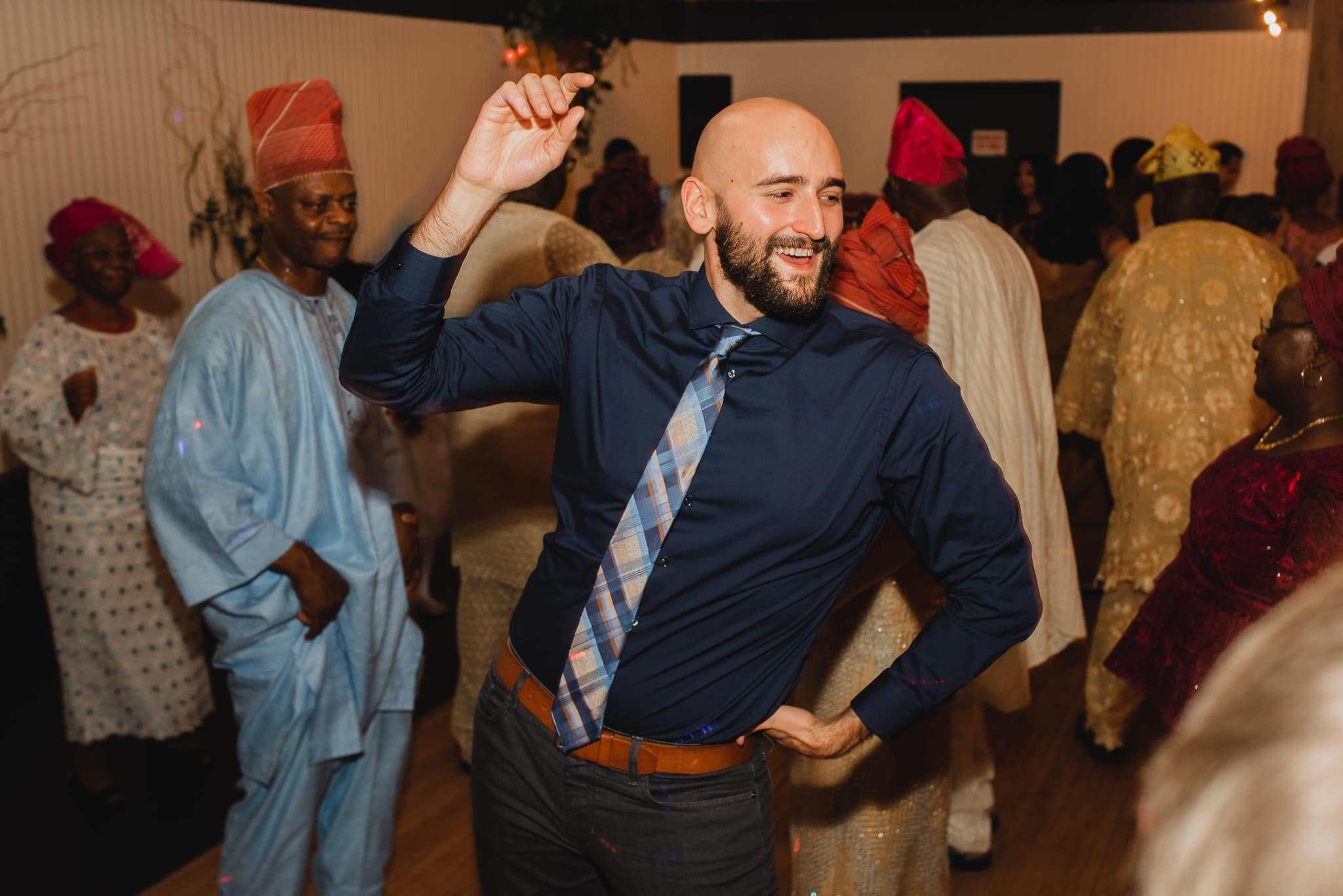 A man in a dark shirt dances joyfully at the lively wedding at the Evergreen, surrounded by people in colorful traditional clothing.