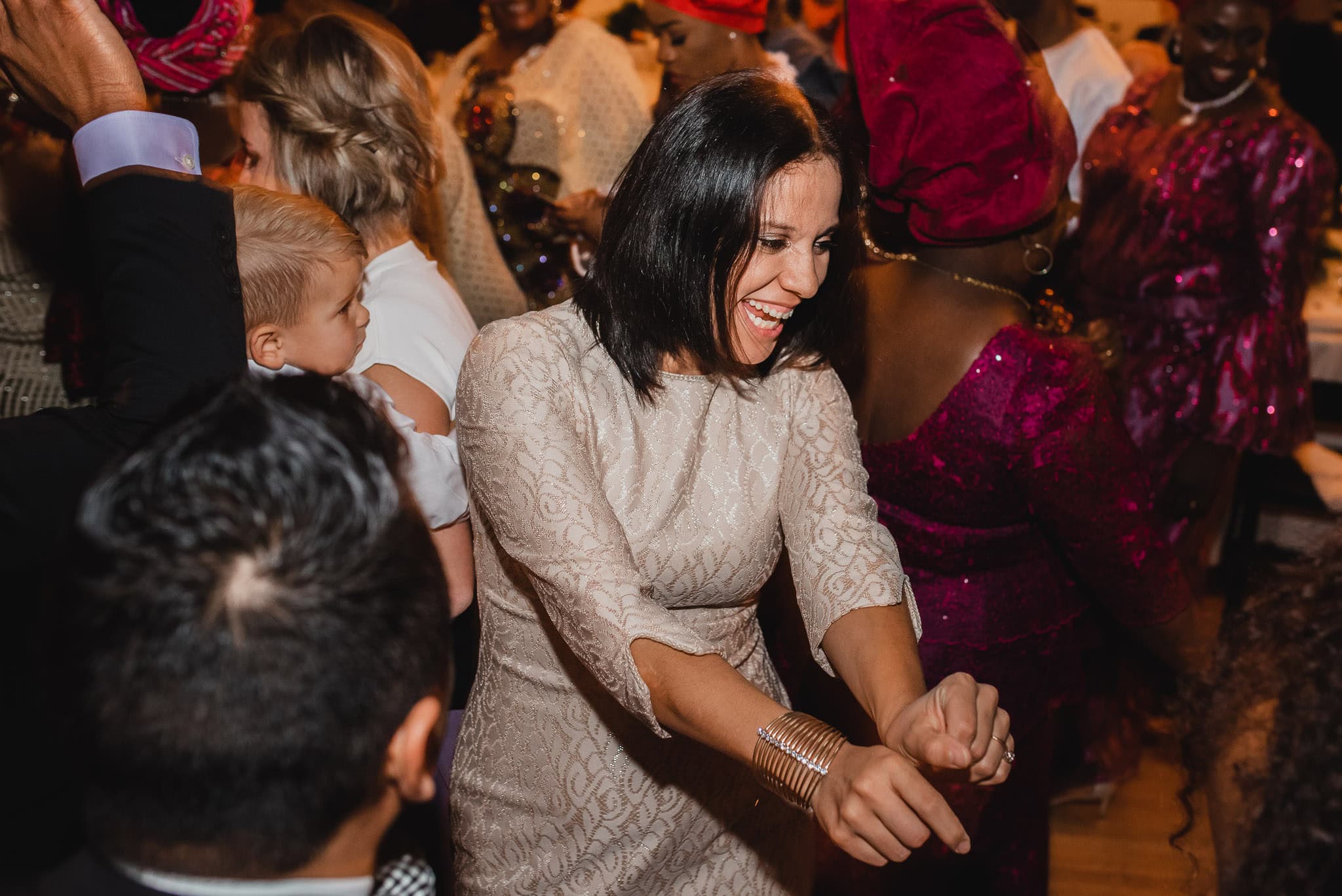 At a wedding at the Evergreen, a woman in a beige dress joyfully dances amid guests in vibrant outfits, creating an atmosphere of sheer celebration.