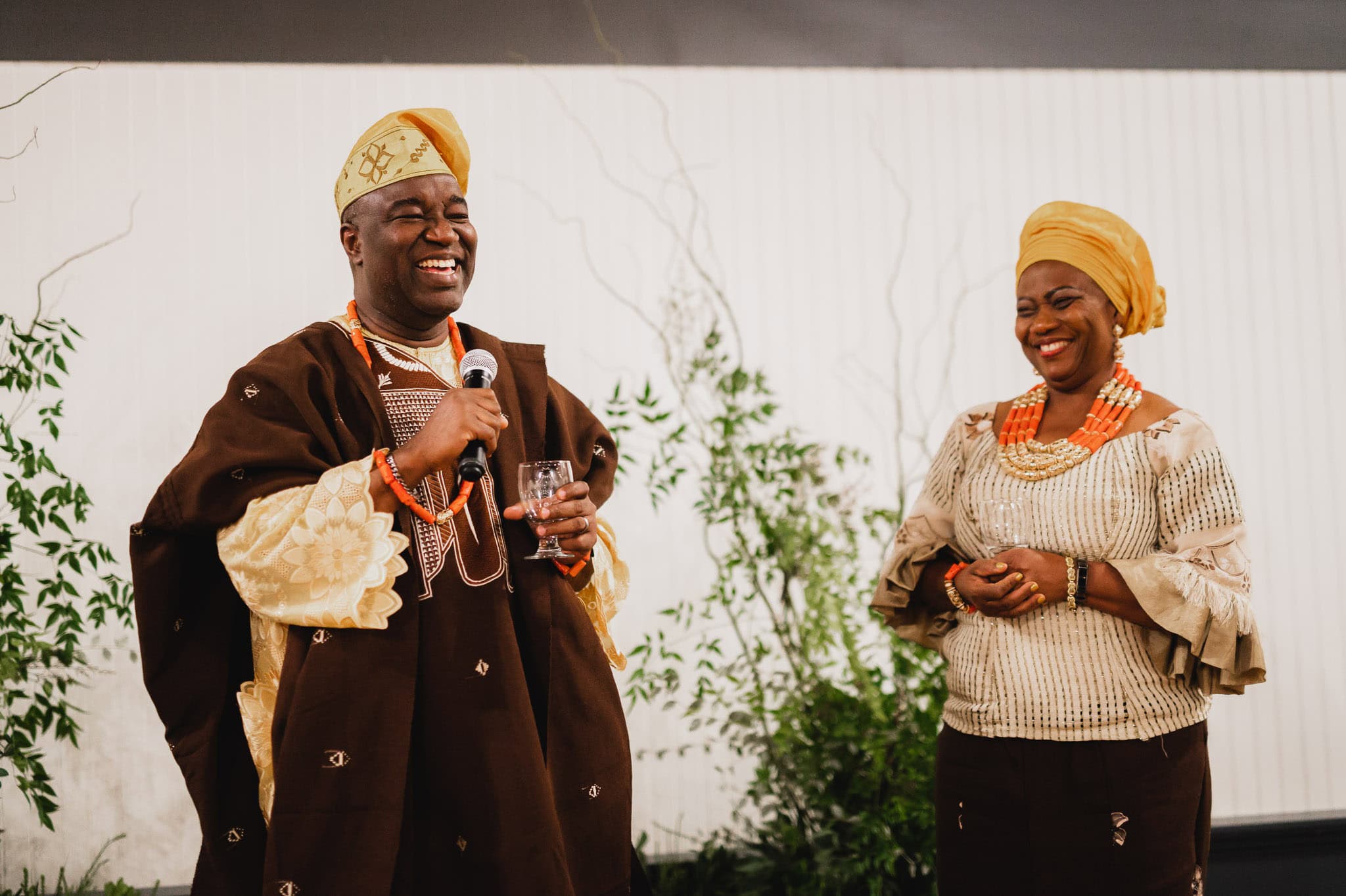 A man and woman in traditional attire smiling; the man holds a microphone and a glass at a wedding at the Evergreen.