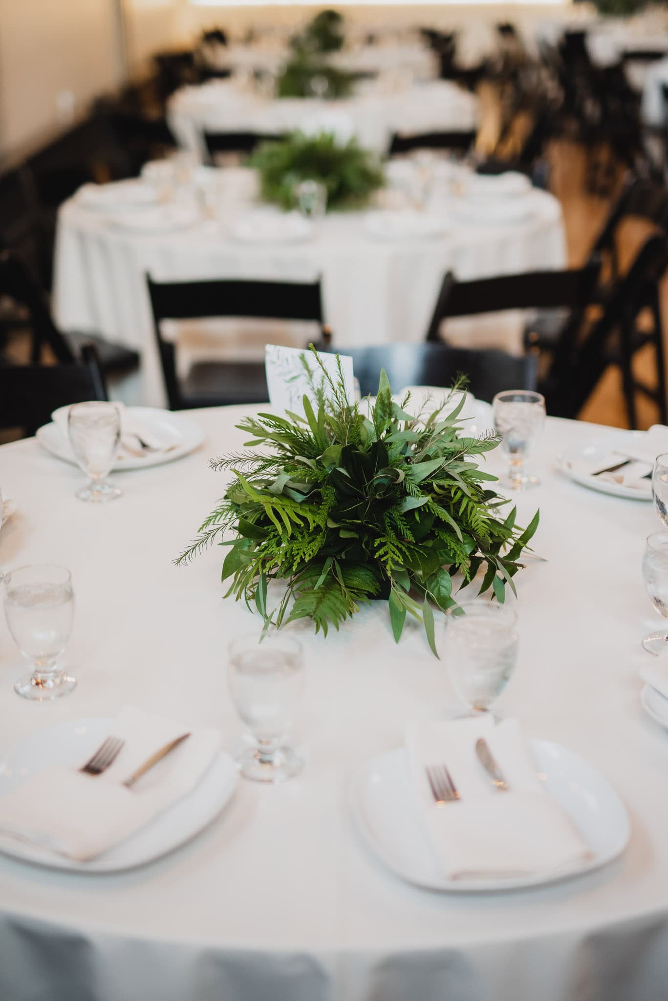 White table settings featuring a greenery centerpiece adorn the round tables, complemented by black chairs and elegant glassware. This sophisticated arrangement creates a perfect setting for a wedding at the Evergreen event space.
