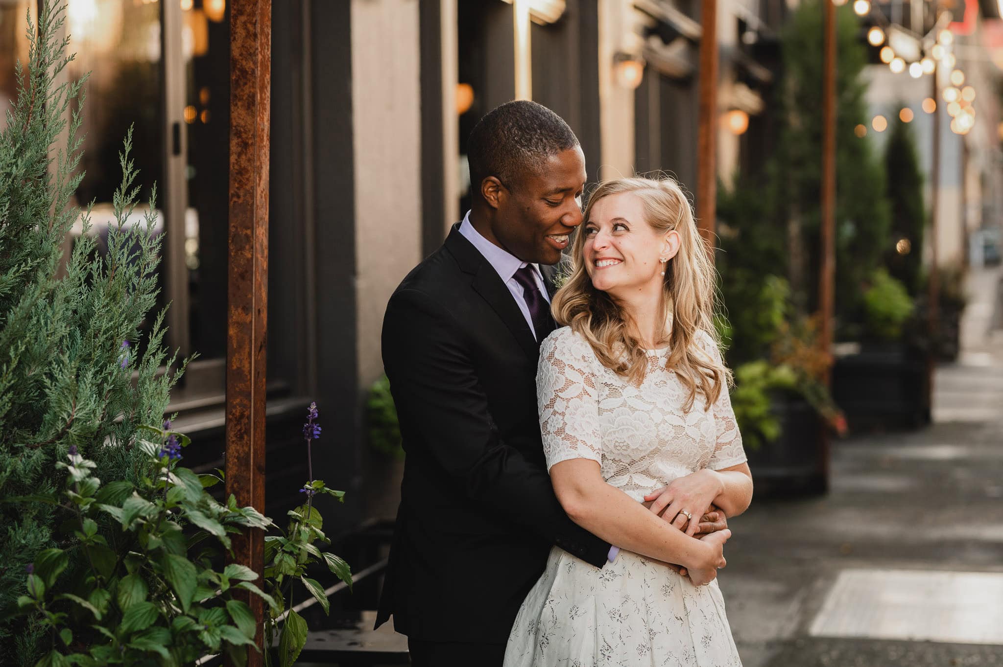A couple embraces and smiles at each other on a city sidewalk lined with plants and lights, celebrating their wedding at the Evergreen.