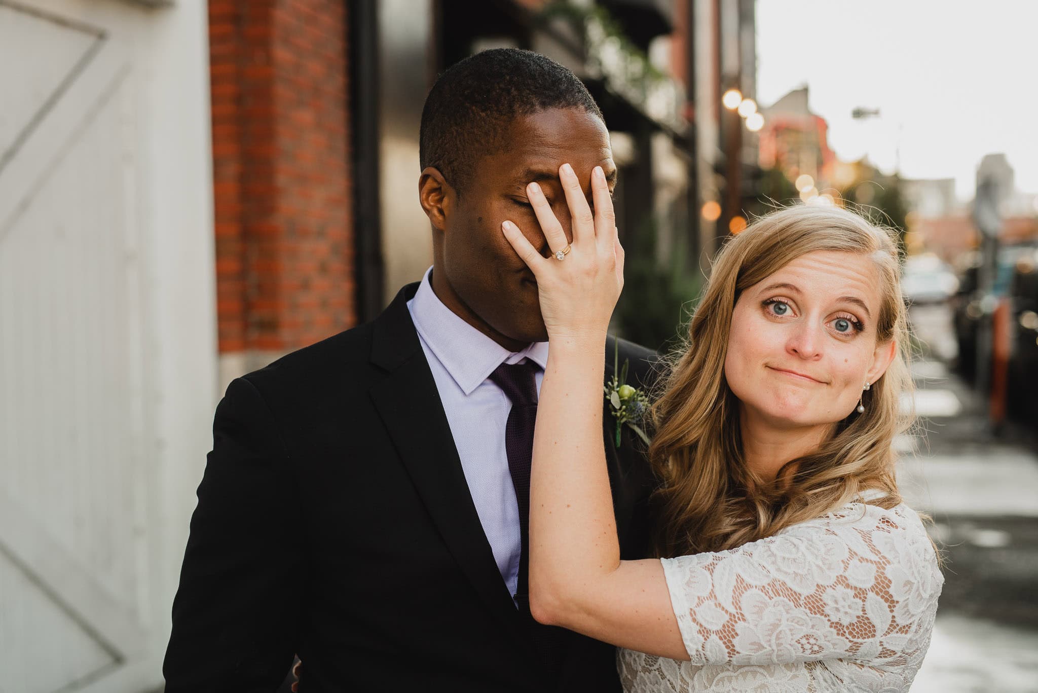 The bride playfully covers the grooms face with her hand while smiling; they stand together on a city street, capturing a joyful moment from their wedding at the Evergreen.