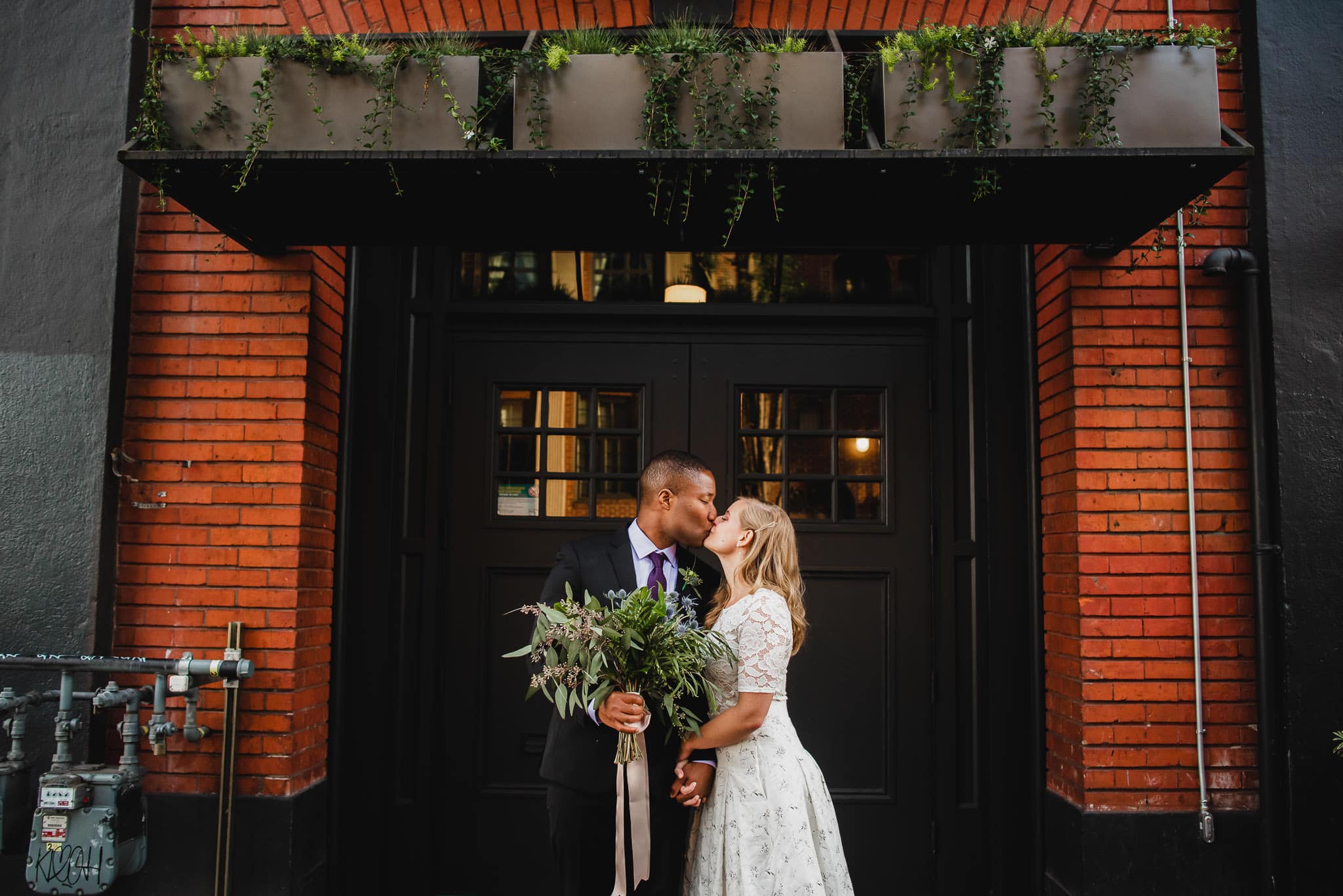 A couple shares a kiss in front of a brick building adorned with plants above, holding a bouquet, creating a picture-perfect moment at their wedding at the Evergreen.
