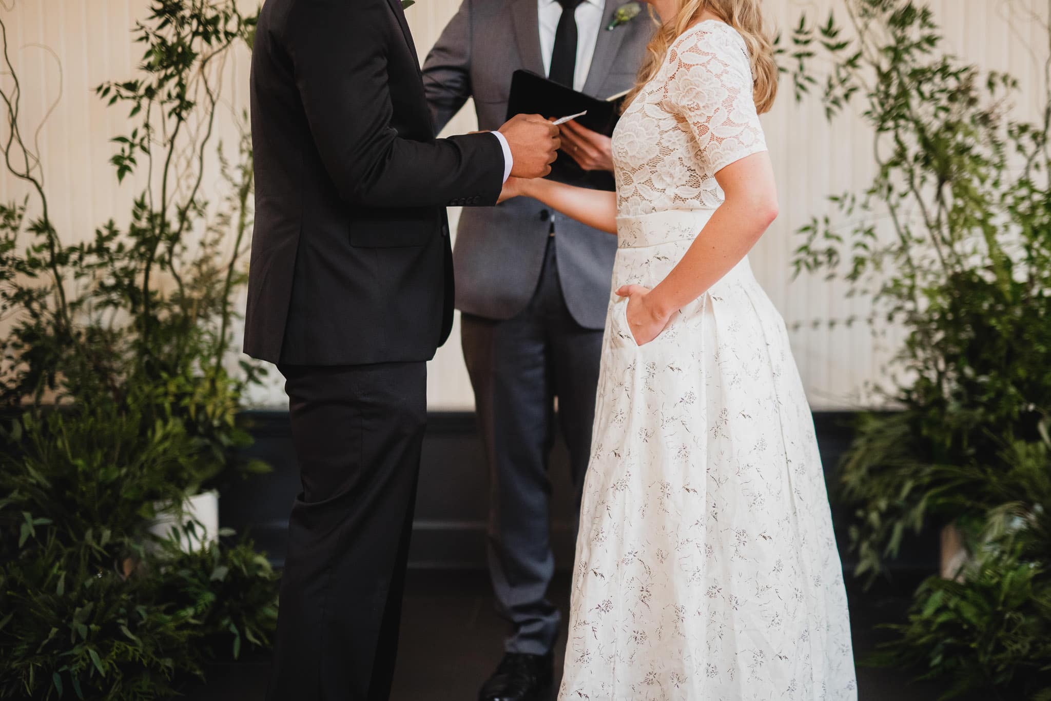 A couple exchanging vows at the wedding at the Evergreen, with a person officiating, surrounded by lush greenery.