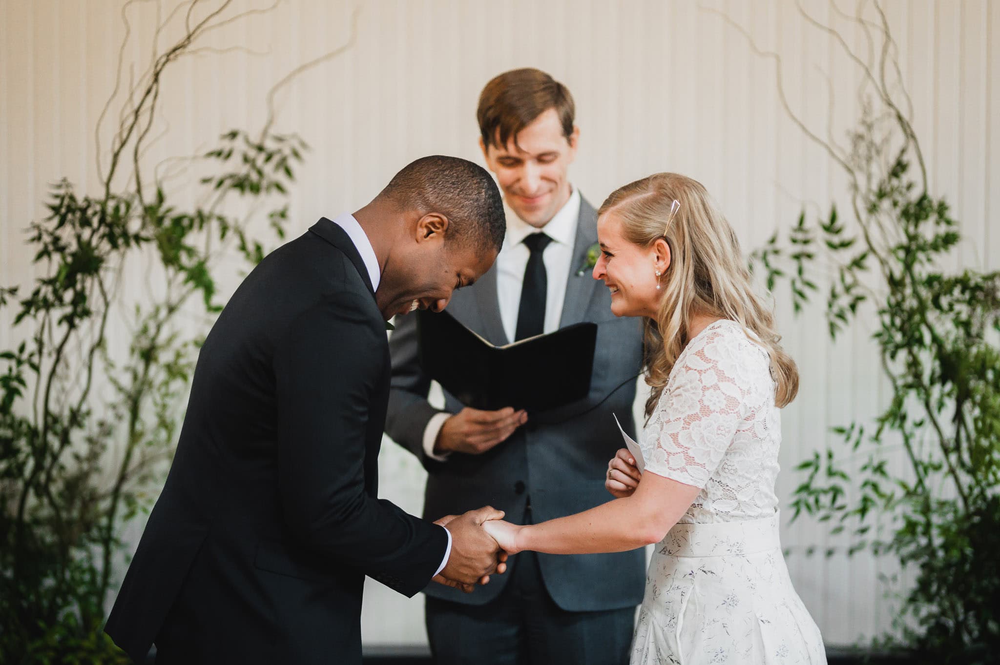 A couple holds hands and smiles warmly during a wedding at the Evergreen, with the officiant standing in the background.