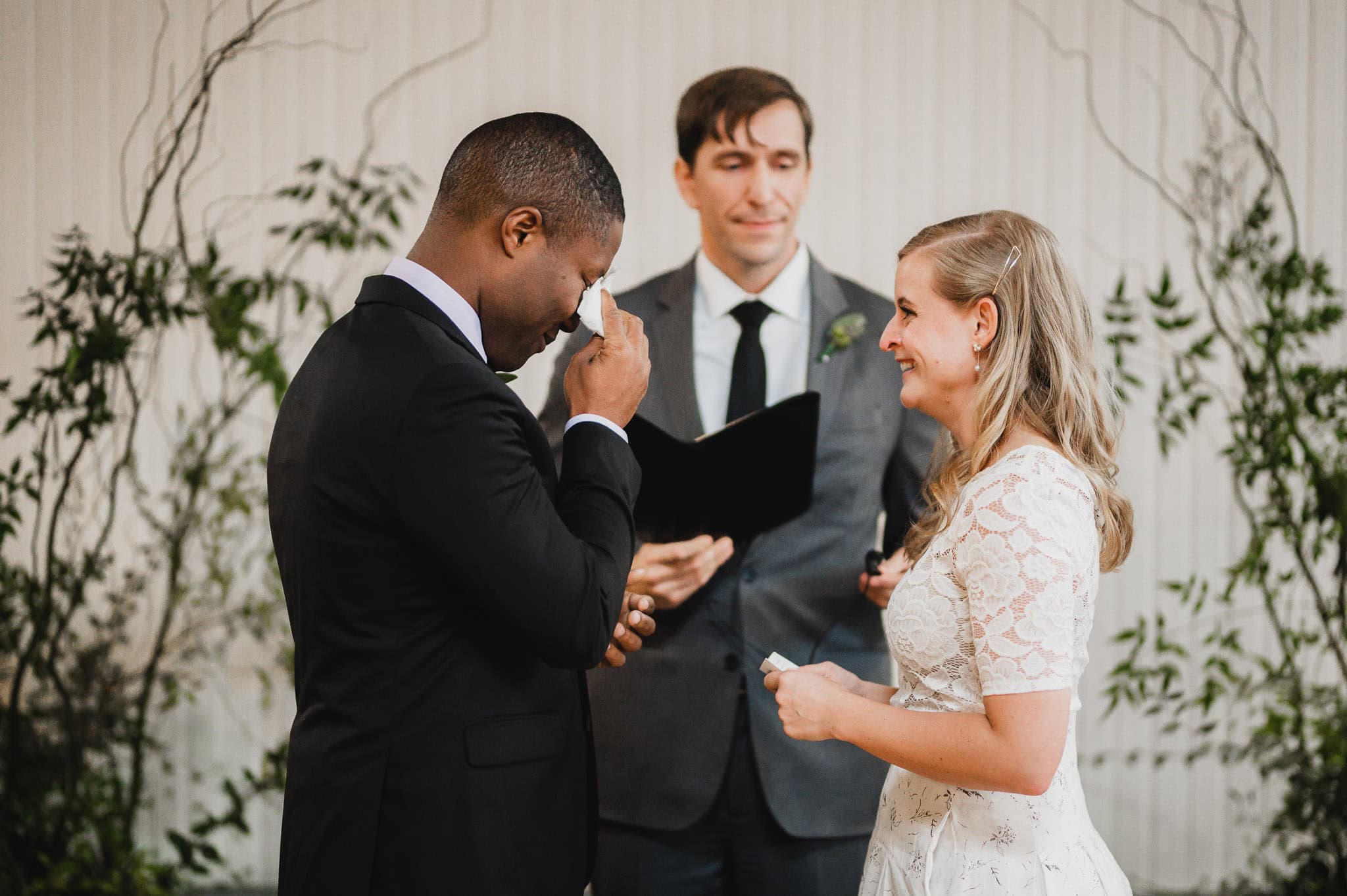 At the wedding at the Evergreen, the bride and groom exchanged vows as the groom wiped tears from his eyes, while the officiant held a book.