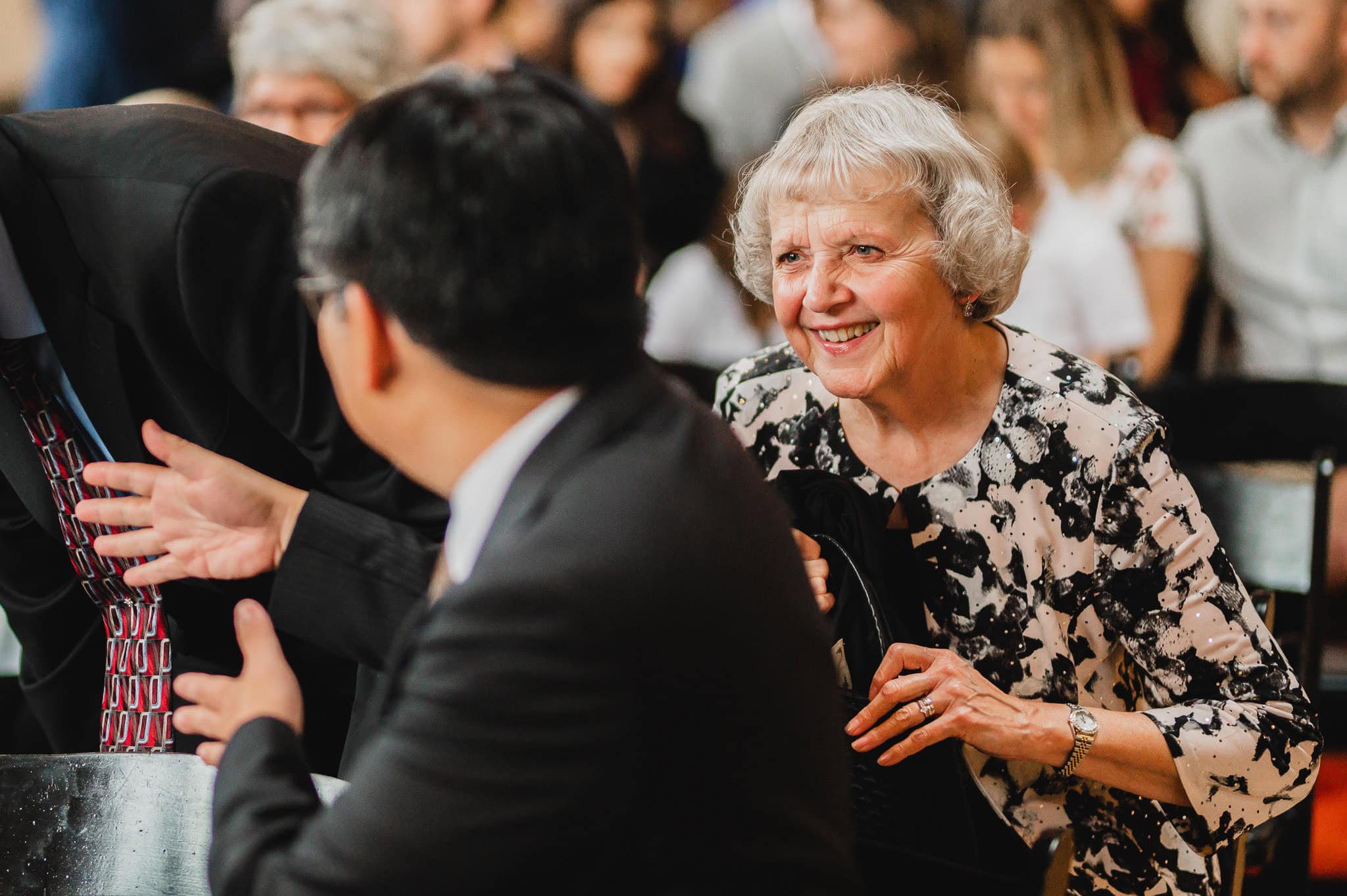 An elderly woman smiles warmly as she engages in conversation at a formal wedding at the Evergreen, with other attendees softly blurred in the background.