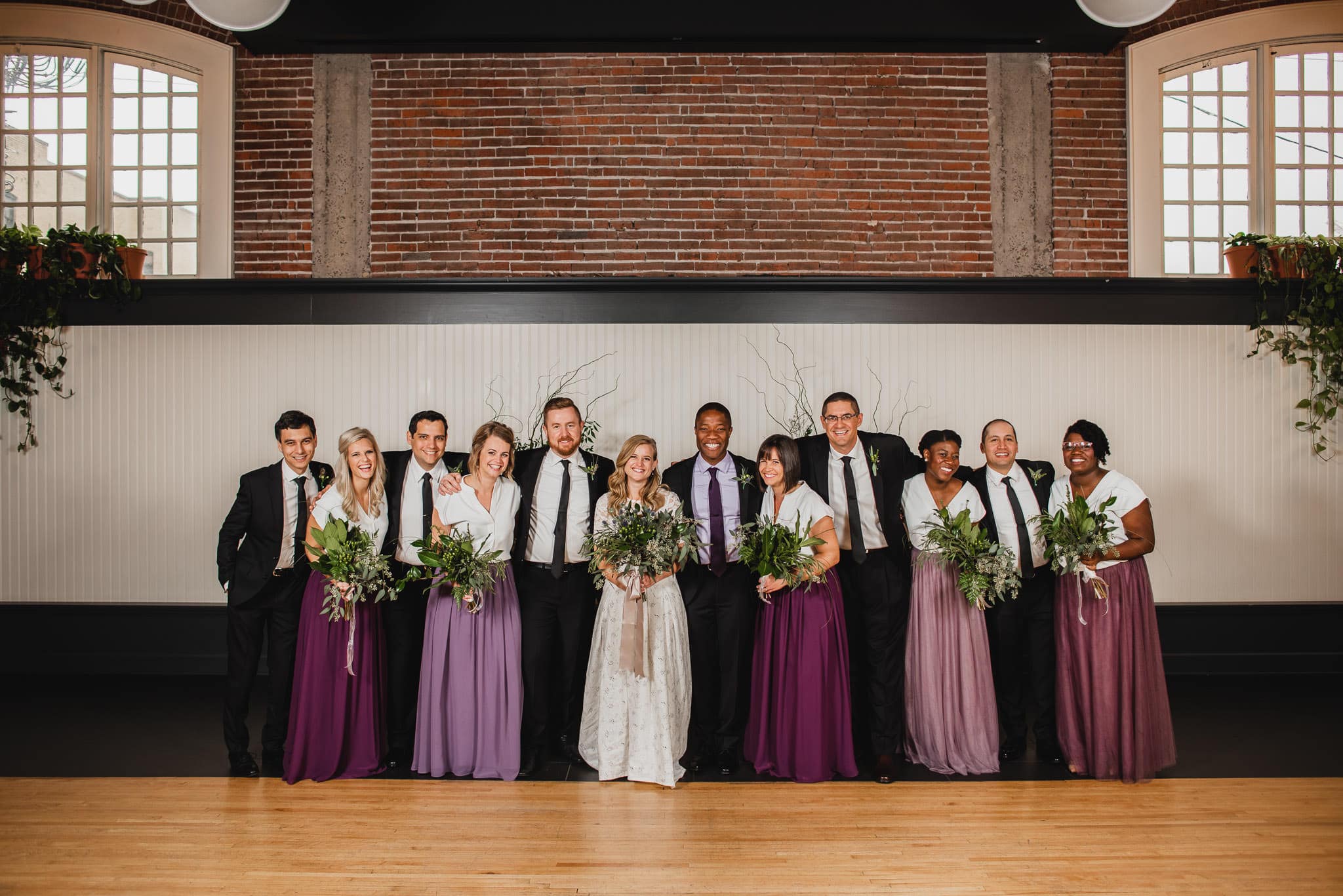 Wedding at the Evergreen featuring the bride, groom, and attendants in purple dresses and black suits, posing elegantly in a brick-walled room.