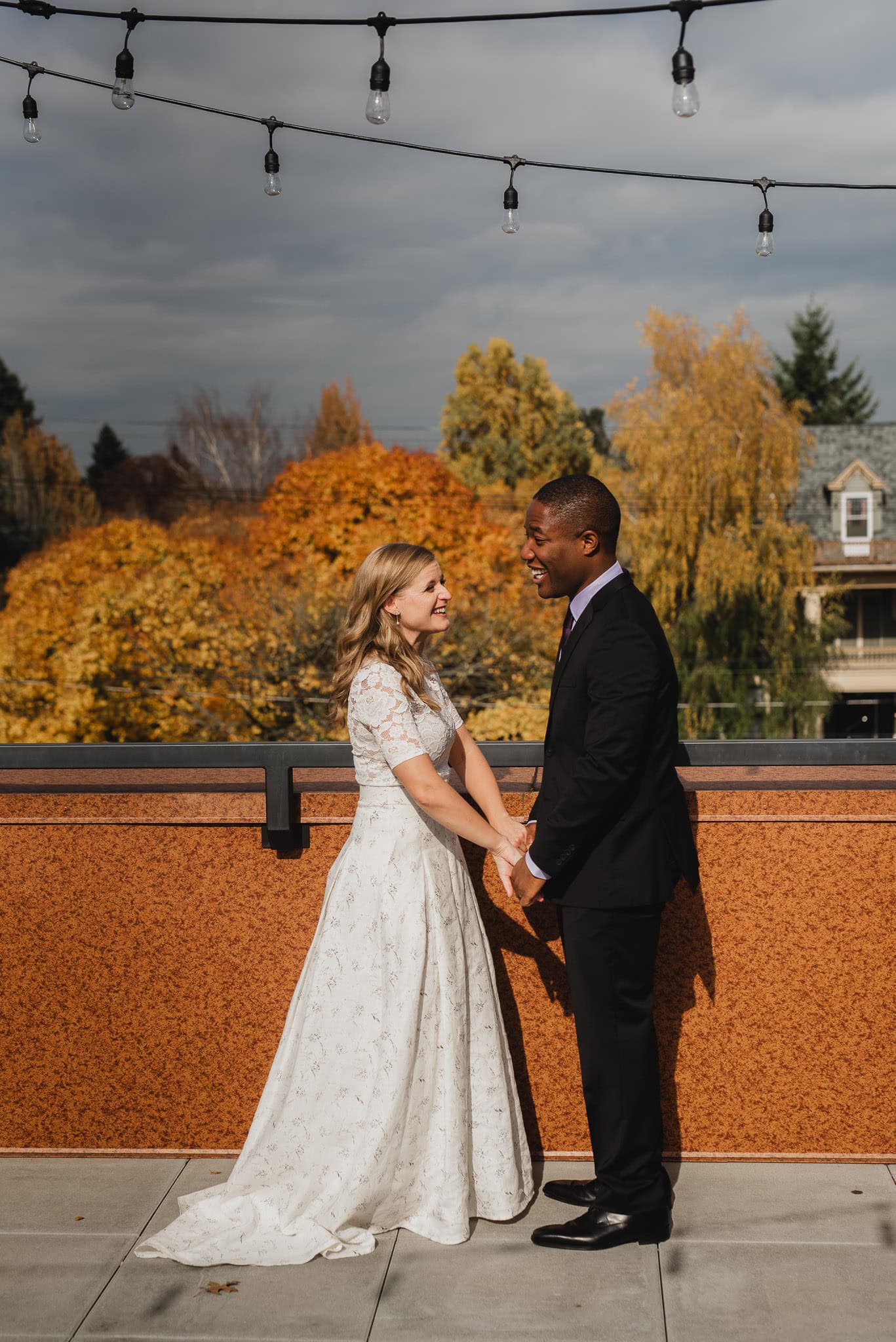 The bride and groom savor their first look on the rooftop of an Airbnb, holding hands against a backdrop of vibrant autumn trees, as they prepare for a wedding at The Evergreen.