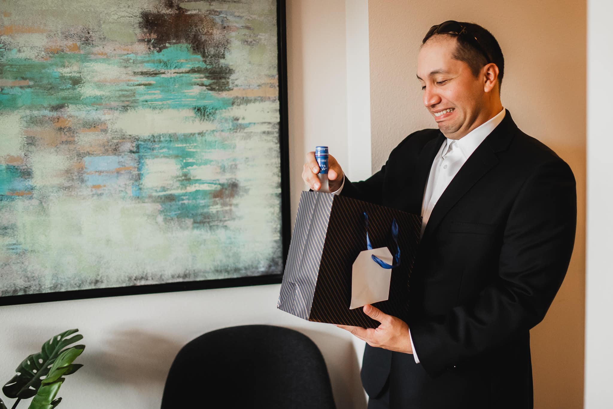 Man in a suit reacts happily to a gift bag, holding a bottle, at an Airbnb rental while getting ready for a wedding at The Evergreen, near an abstract painting.