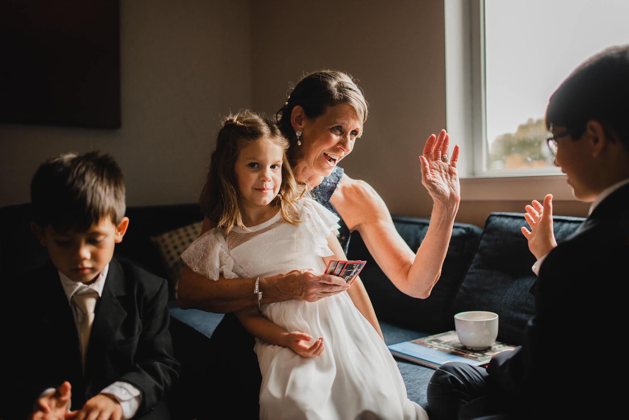 A woman and three children are smiling and playing with cards indoors, near a window on a sunny day, at an Airbnb rental while getting ready for a wedding at The Evergreen.