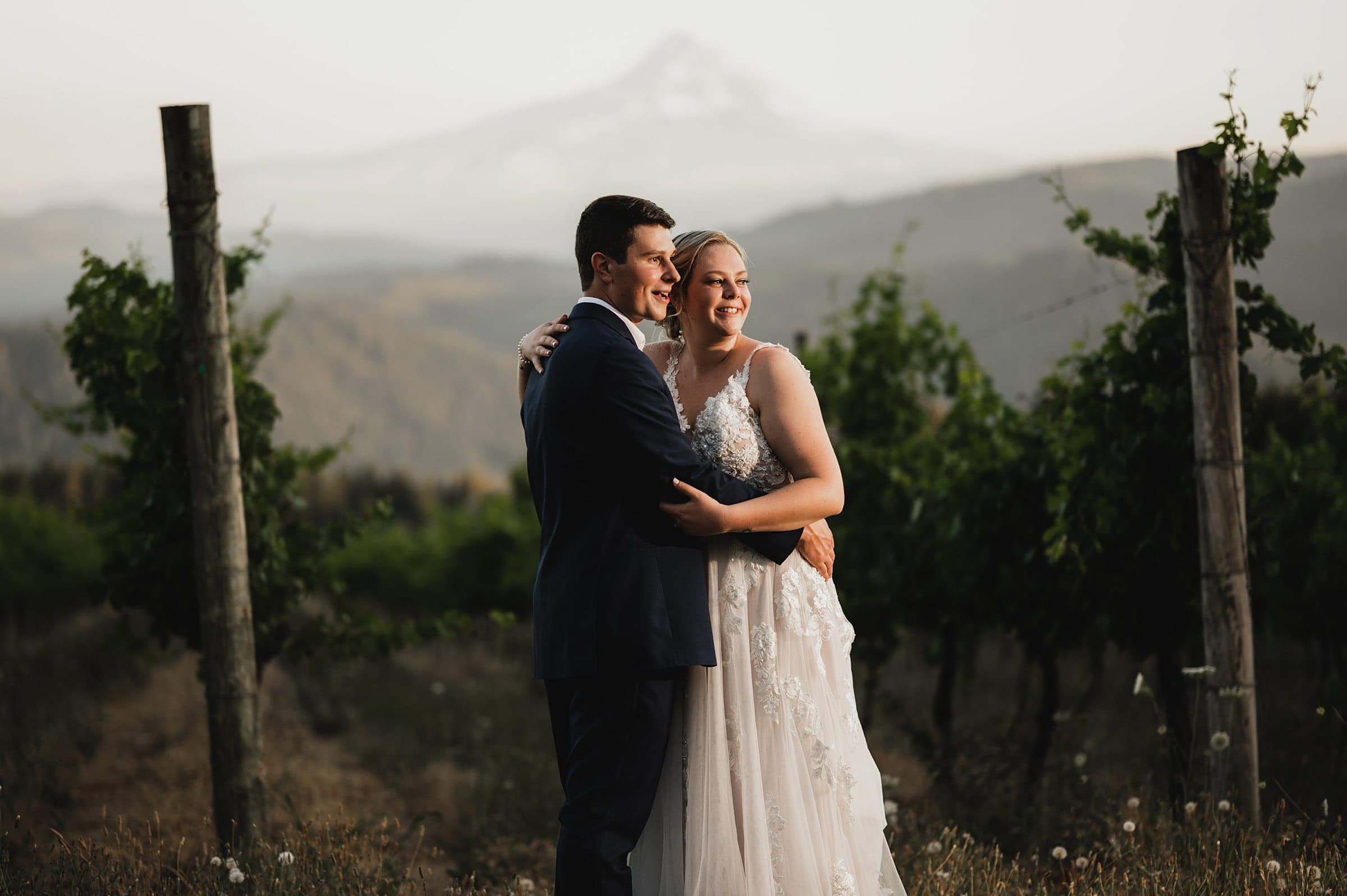 A couple in wedding attire embrace amidst the lush rows of Gorge Crest Vineyards, with majestic Mt Hood towering in the background.