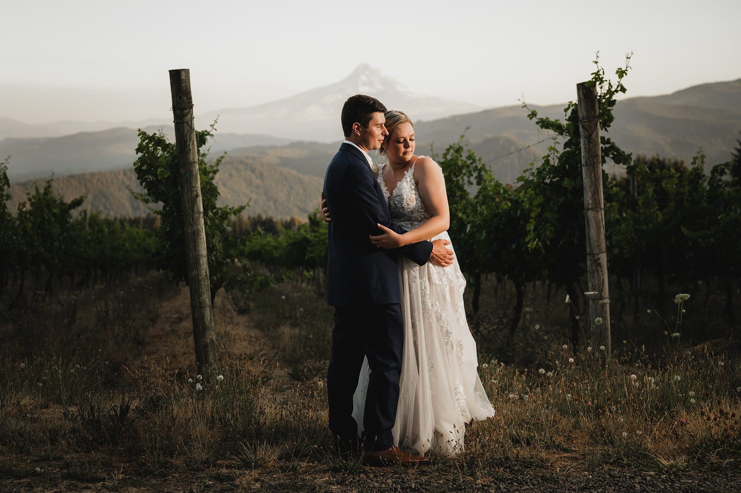 A couple in wedding attire embraces in a vineyard at Gorge Crest, with majestic Mt Hood as the backdrop.