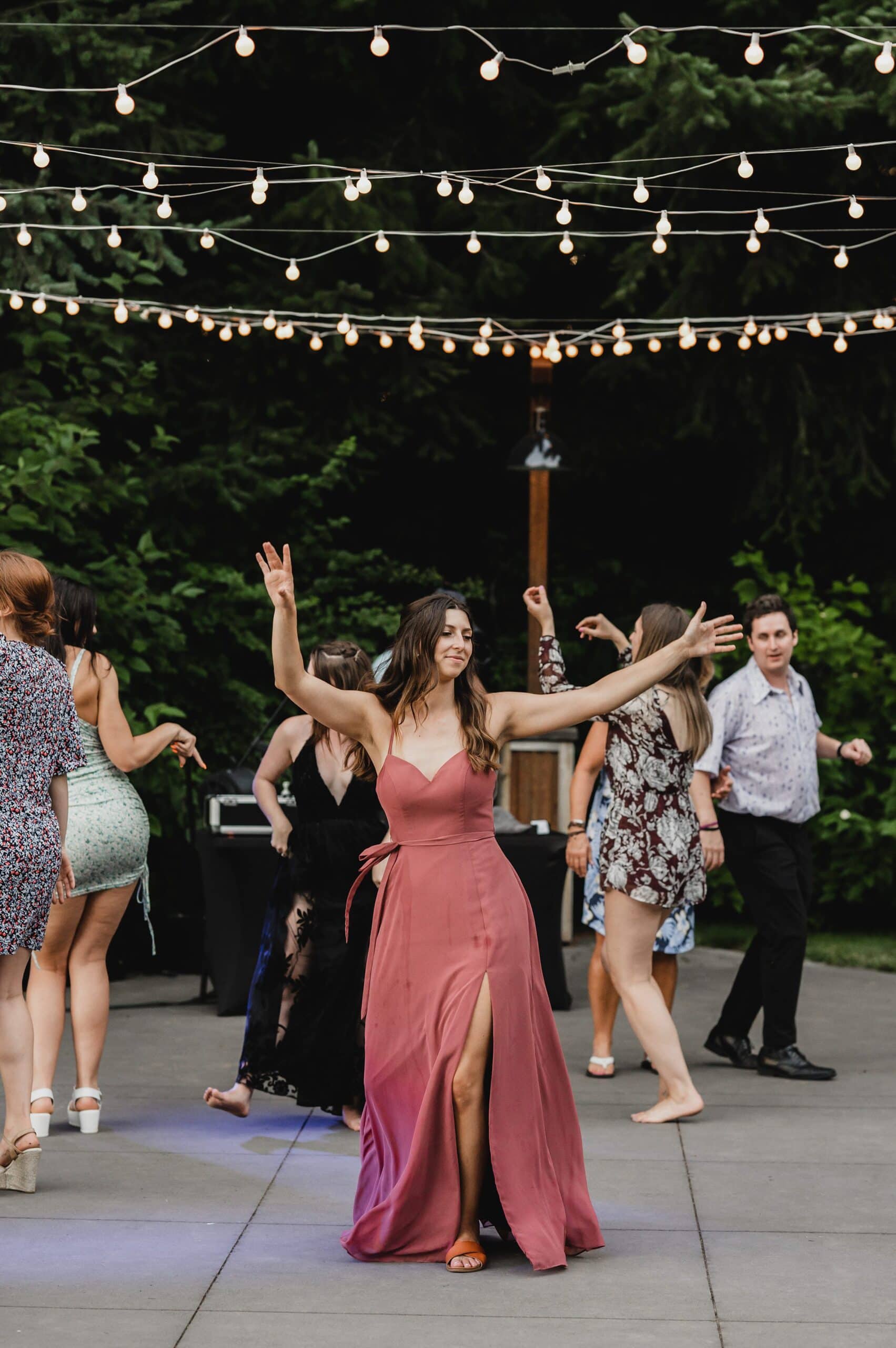 A woman in a pink dress dances joyfully under string lights, surrounded by others at an outdoor event. The scene captures the vibrant energy of wedding photos at Gorge Crest Vineyards.