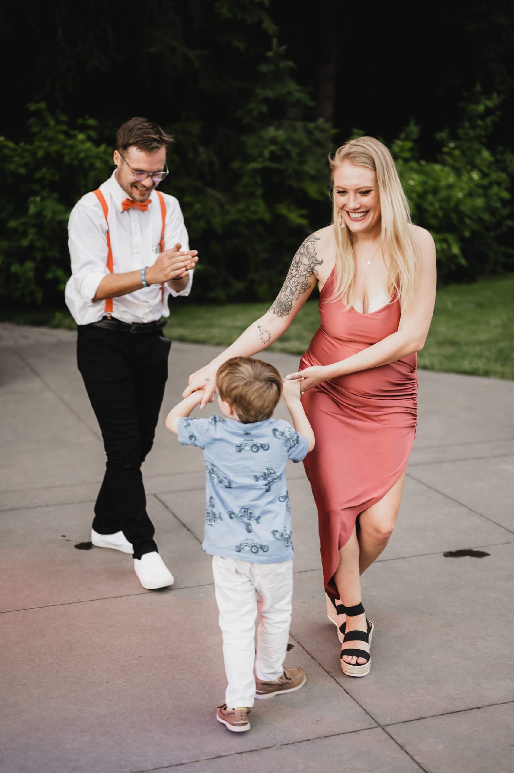 A woman in a pink dress dances with a child on a patio at Gorge Crest Vineyards, surrounded by lush greenery, while a man with suspenders claps nearby.