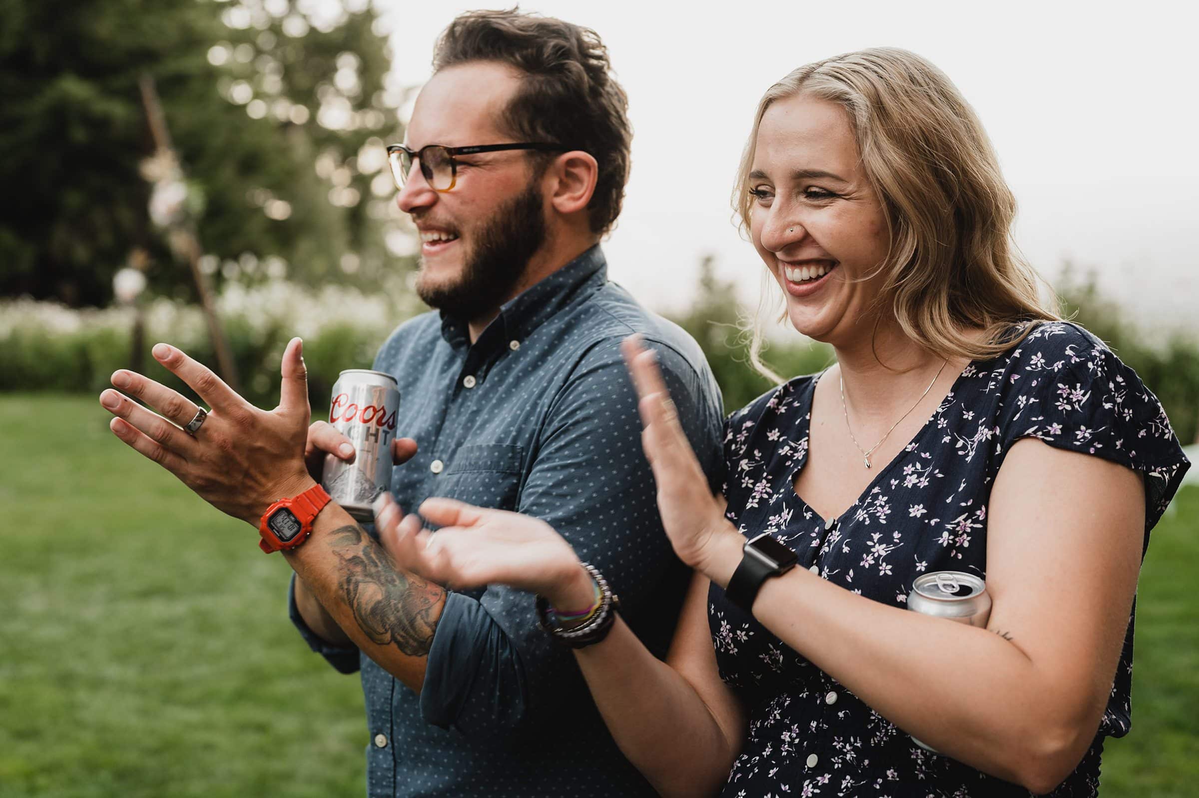 Two people smiling and clapping outdoors, holding beverage cans at a Gorge Crest Vineyard wedding, with greenery in the background.