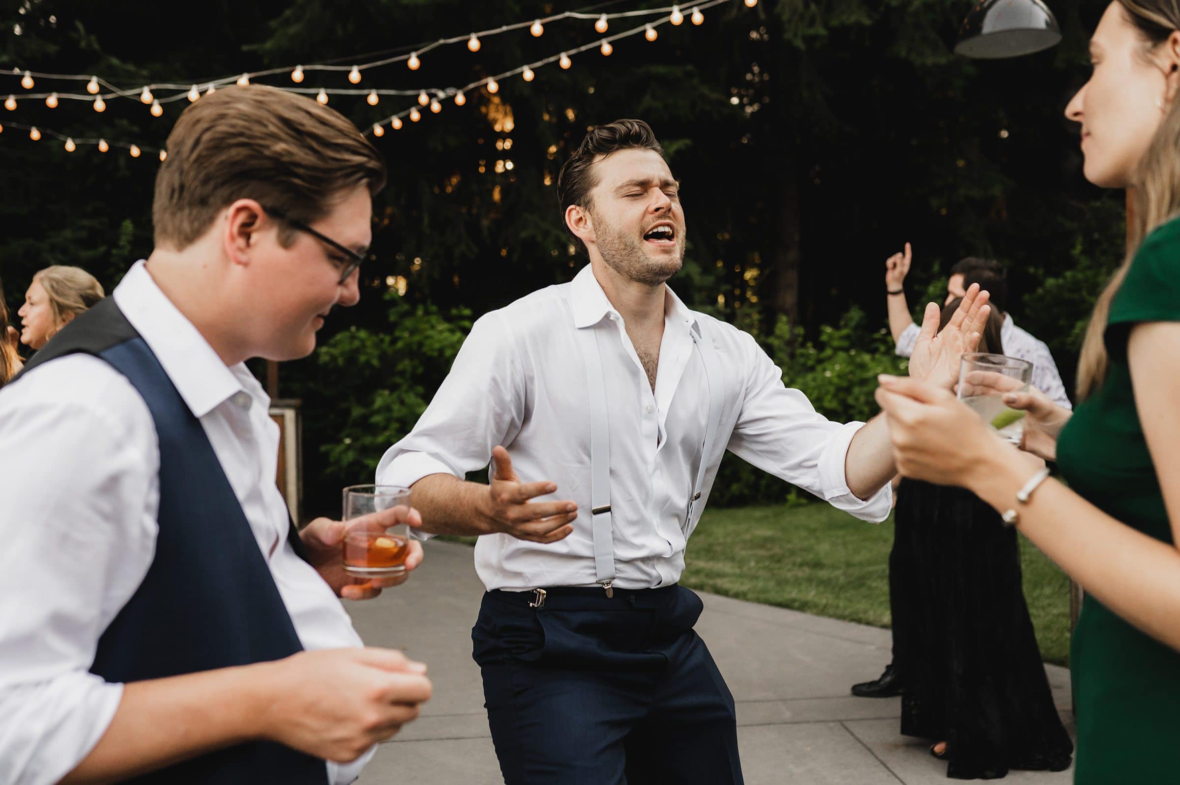 A man in a white shirt is dancing energetically outdoors at a party at Gorge Crest, holding a drink. Other people are in the background, capturing lively moments perfect for wedding photos.