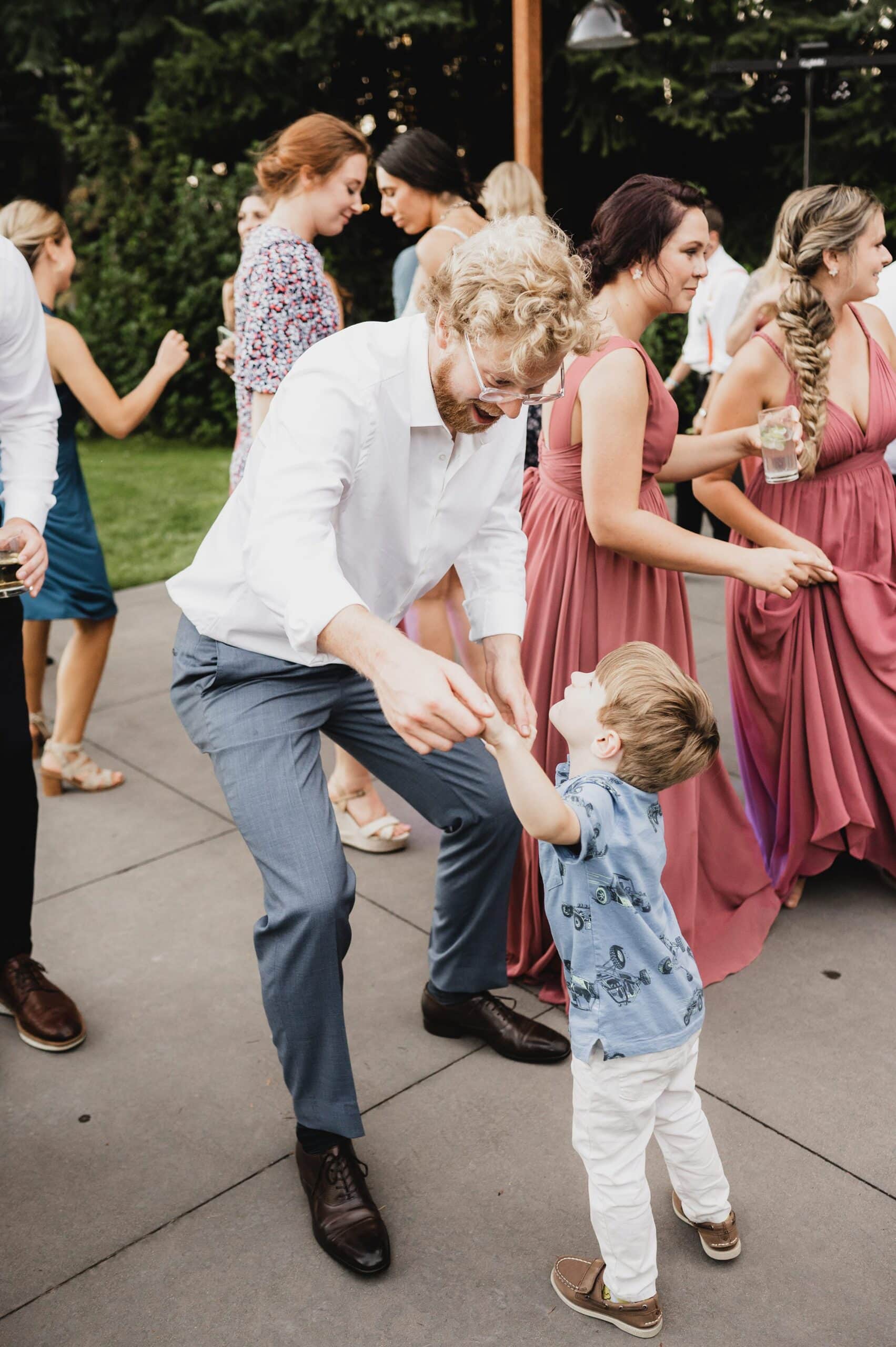 At a lively Gorge Crest Vineyards wedding, a man dances joyfully with a young boy among guests who are also dancing and socializing.