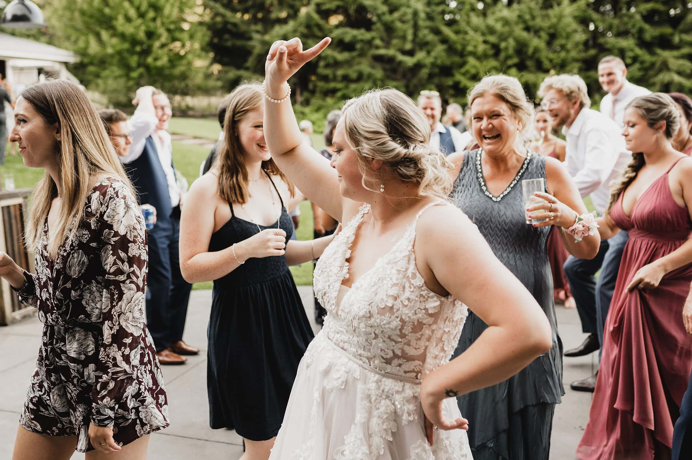 At Gorge Crest Vineyard, a lively group dances joyfully outside at a wedding, led by a woman in a flowing white dress with her arm raised high.