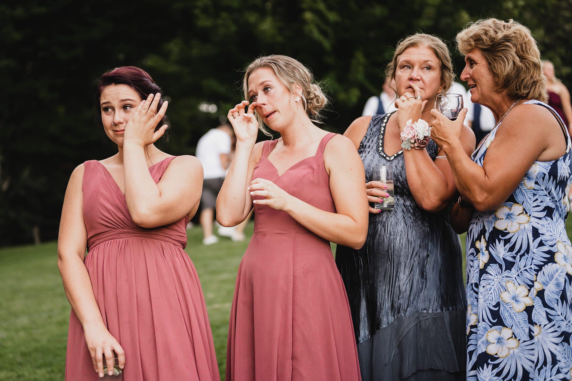 Four women stand outside in formal dresses at a Gorge Crest Vineyards wedding, wiping tears from their eyes during an emotional moment.