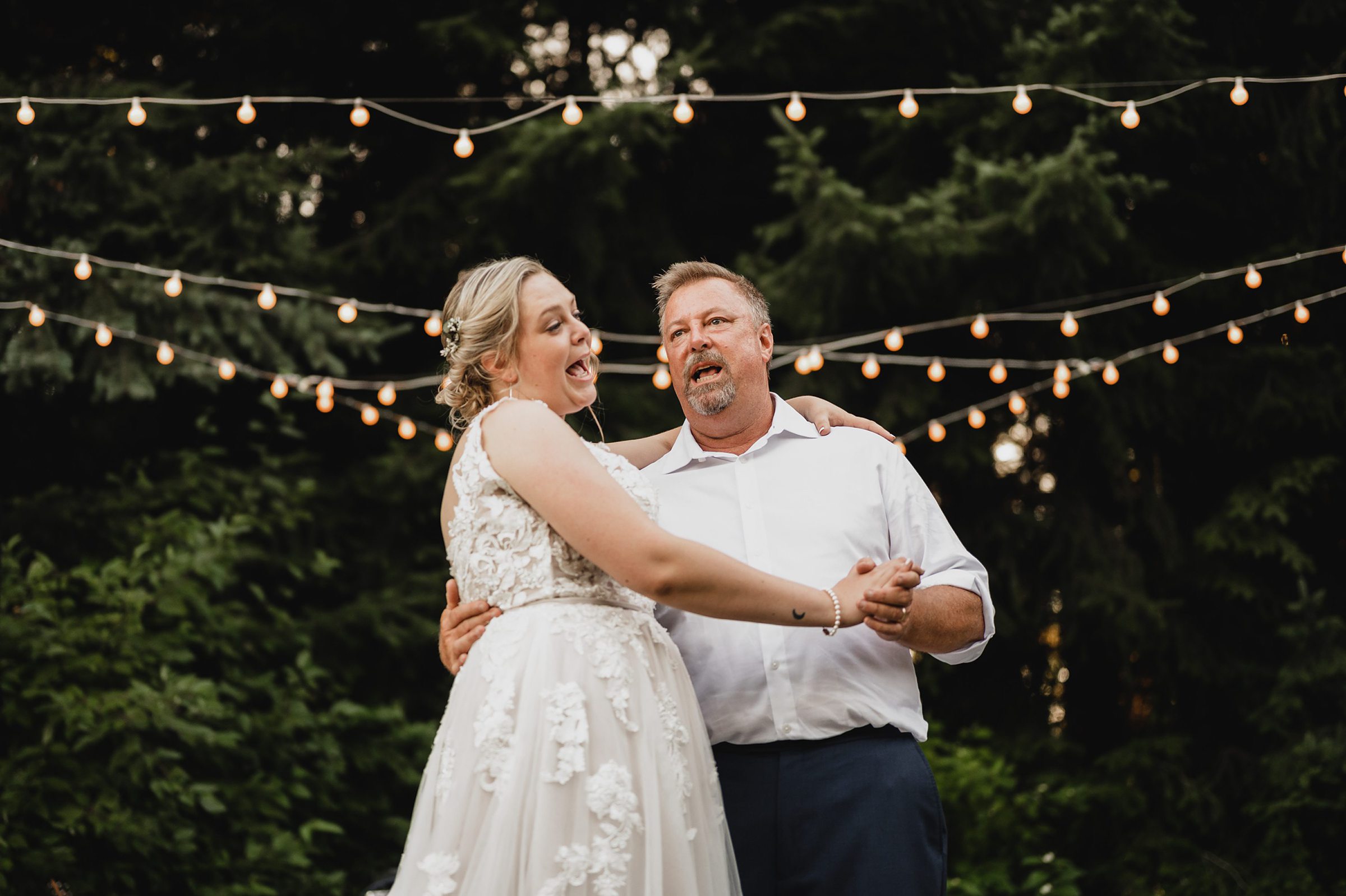 A bride and a man dance under string lights outdoors at dusk, surrounded by trees, during a beautiful Gorge Crest Vineyard wedding.