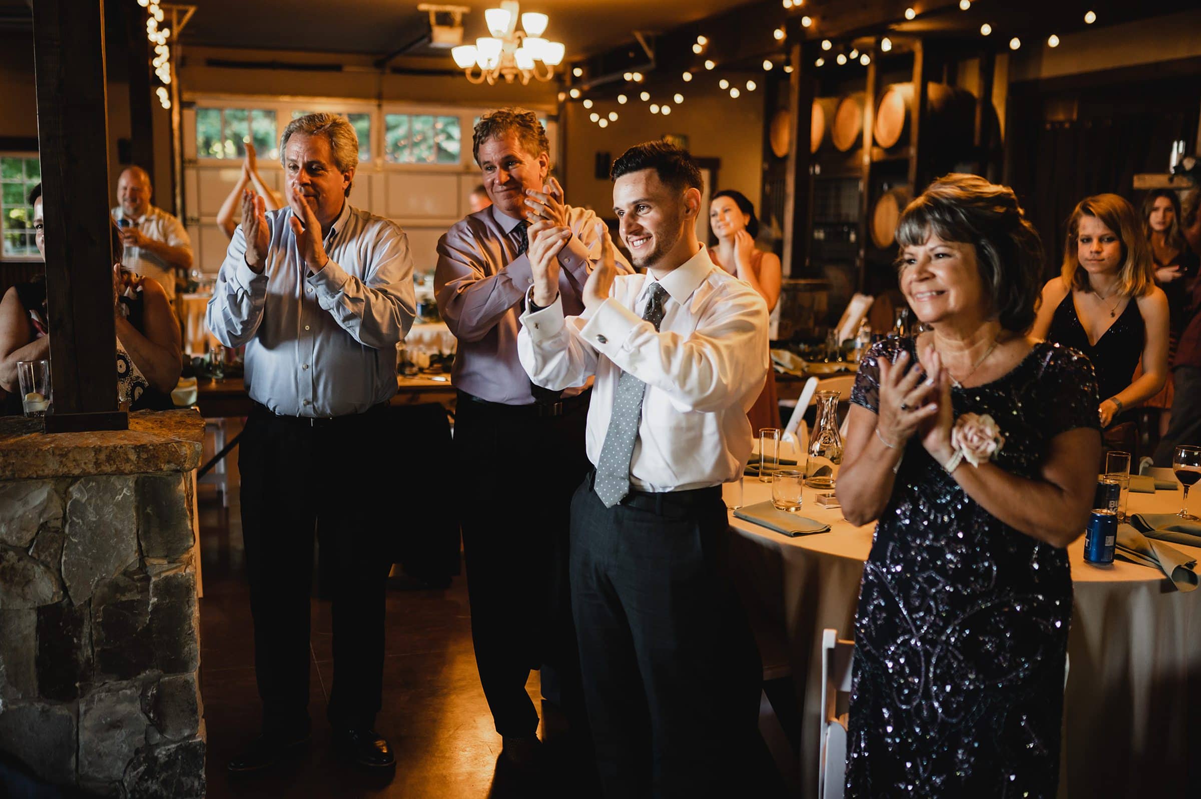 Guests in formal attire clap enthusiastically at an indoor event illuminated by string lights, while others are seated around elegantly decorated tables. The atmosphere echoes a memorable celebration, capturing the spirit of a Gorge Crest Vineyard wedding.
