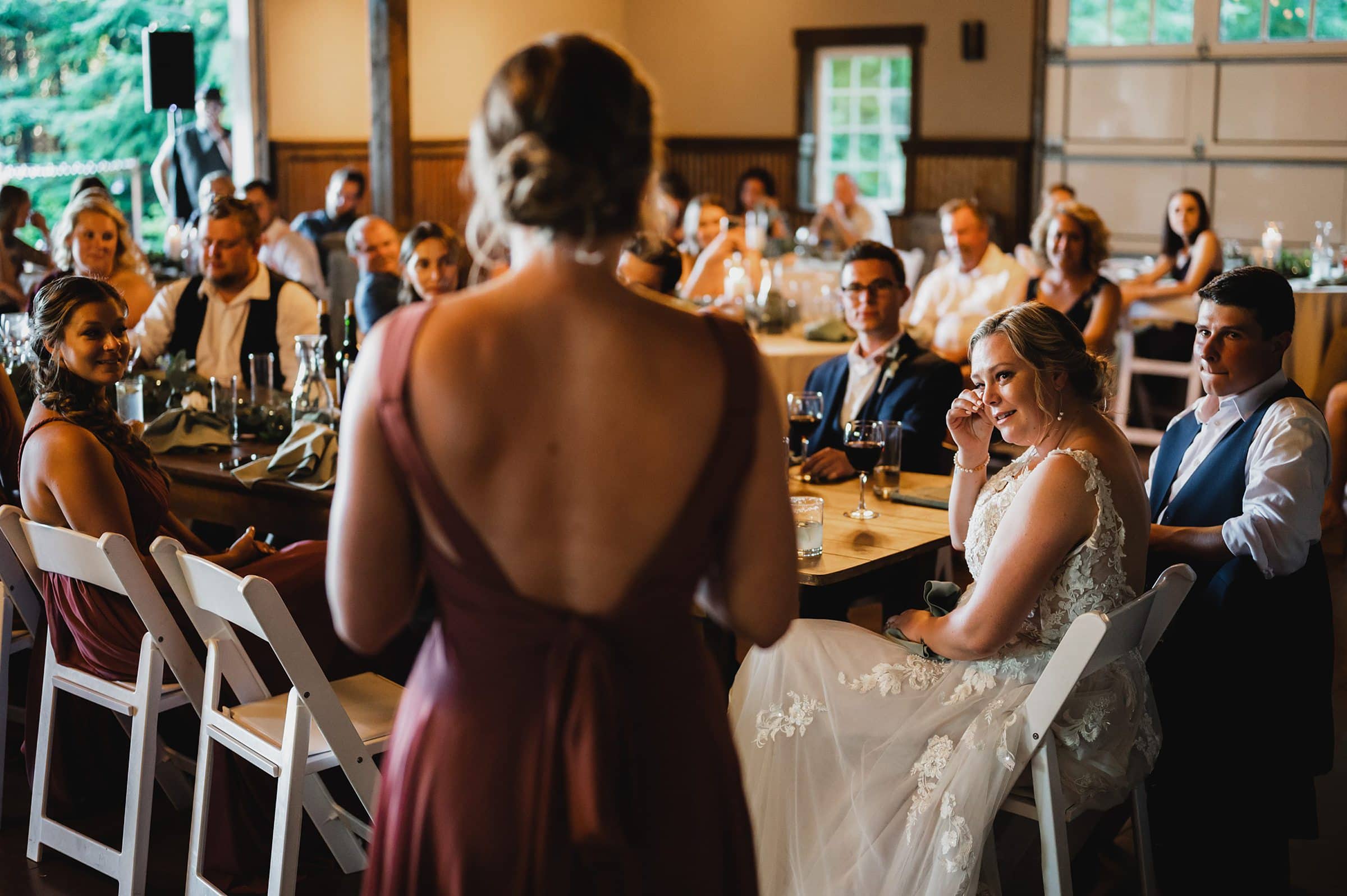 A woman in a brown dress gives a speech at a Gorge Crest Vineyard wedding reception, as guests, including the bride, listen attentively.
