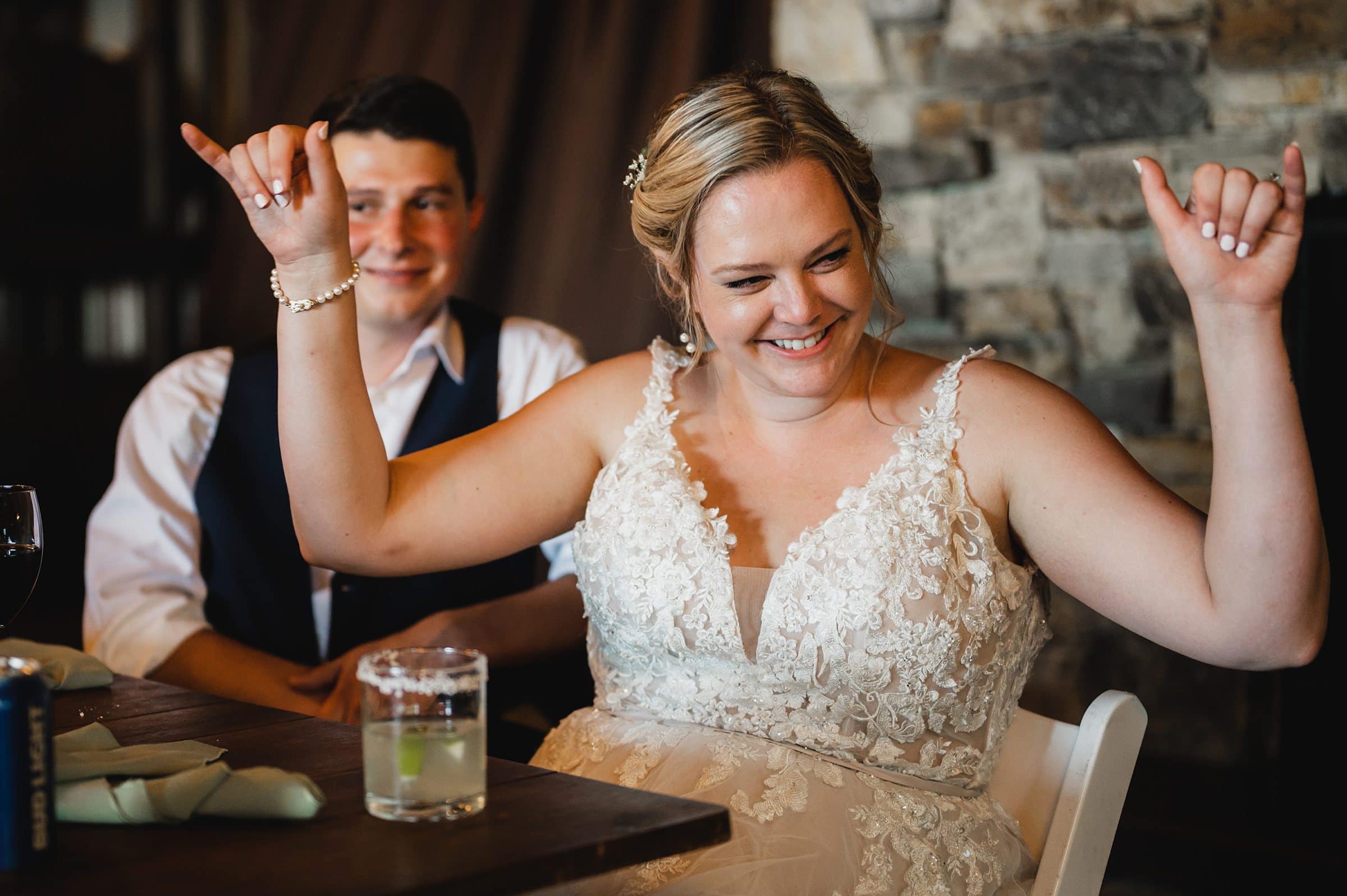 The bride beams as she raises her arms in celebration, with a man in a vest sitting beside her during their Gorge Crest Vineyard wedding.