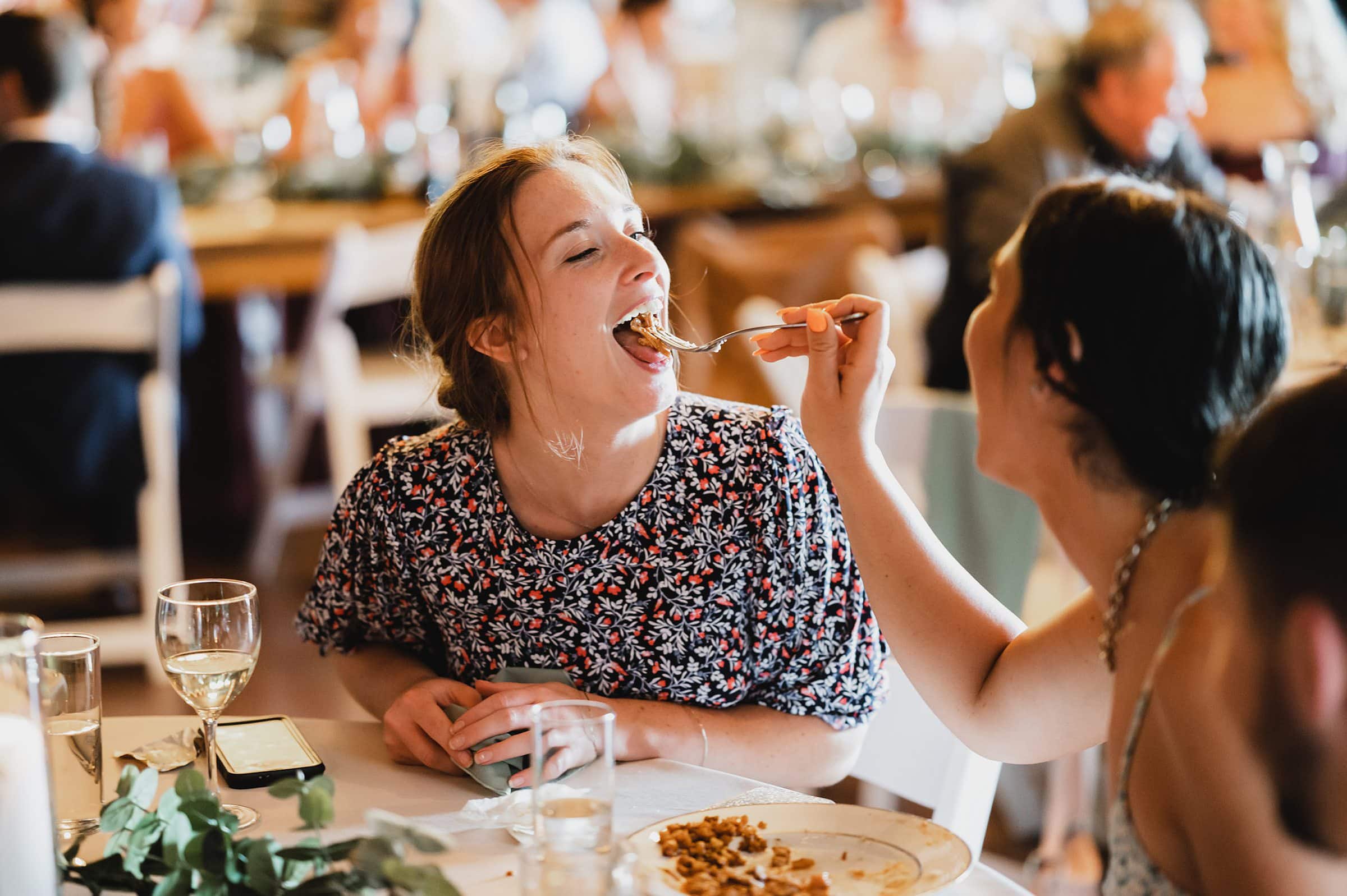 At a table surrounded by drinks and a plate of food, two people smile as one feeds the other. Capturing this candid moment feels like a perfect snapshot from Gorge Crest Vineyards wedding photos.