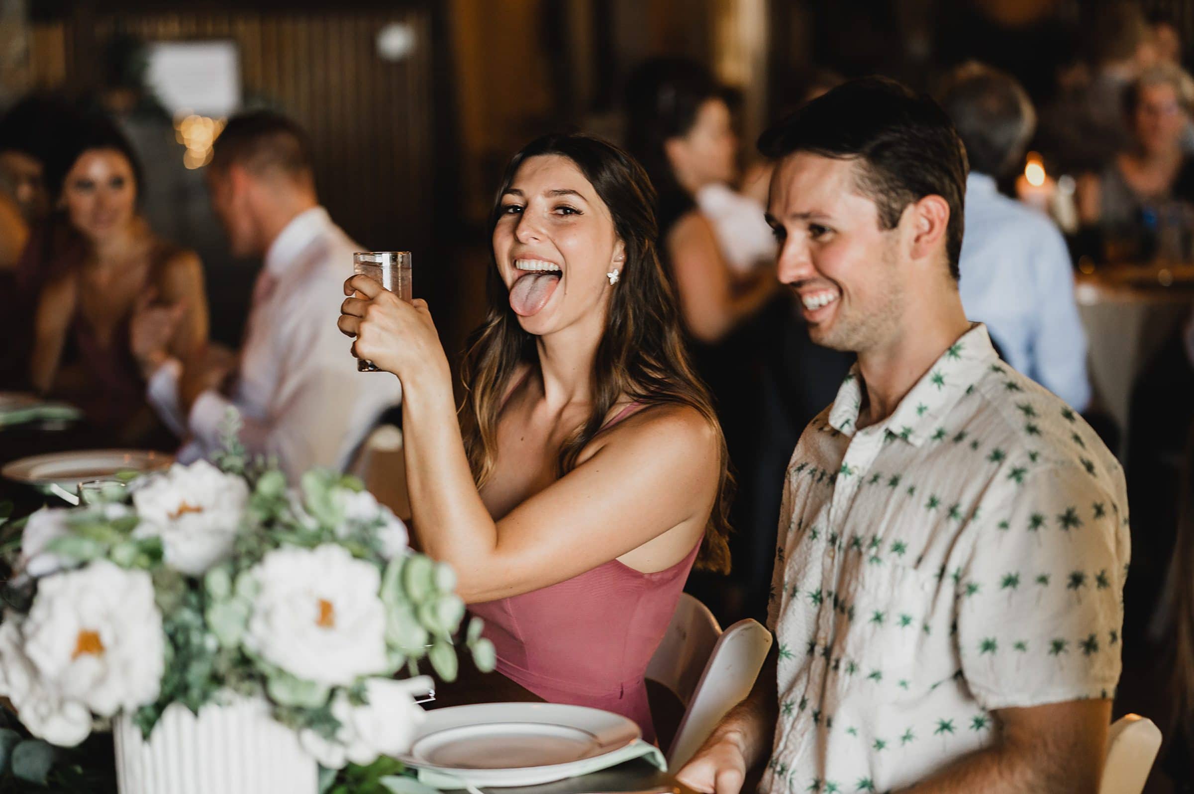 At a festive gathering, a woman in a pink dress playfully sticks her tongue out while holding a drink next to a smiling man. They both seem to be enjoying the joyous atmosphere, perhaps captured amidst wedding photos at Gorge Crest Vineyard.
