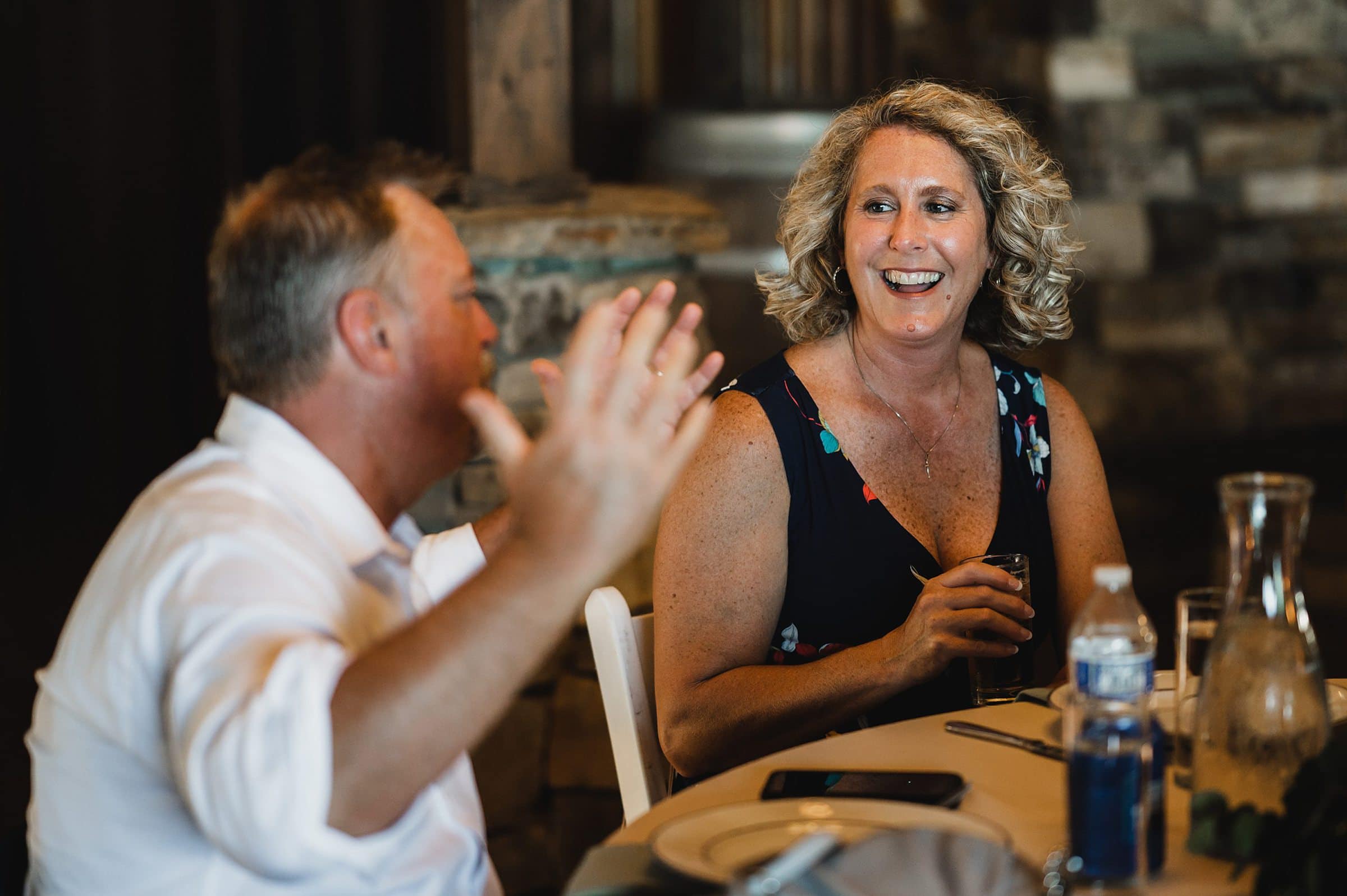Two people are sitting at a table, talking and smiling at a Gorge Crest Vineyard wedding. One gestures animatedly, while the other holds a drink.