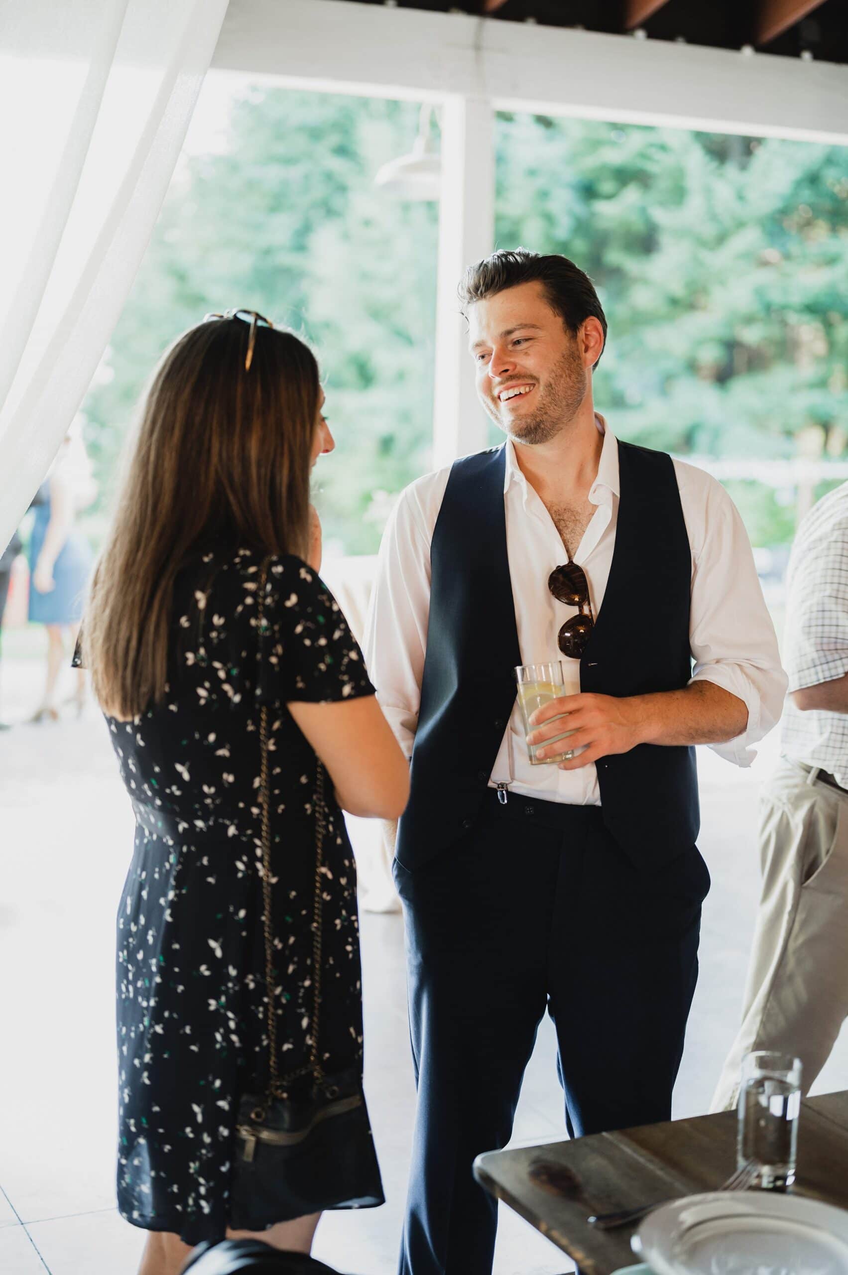 A man and woman stand indoors at a Gorge Crest wedding, engaging in conversation. The man holds a drink, and they both look pleased.
