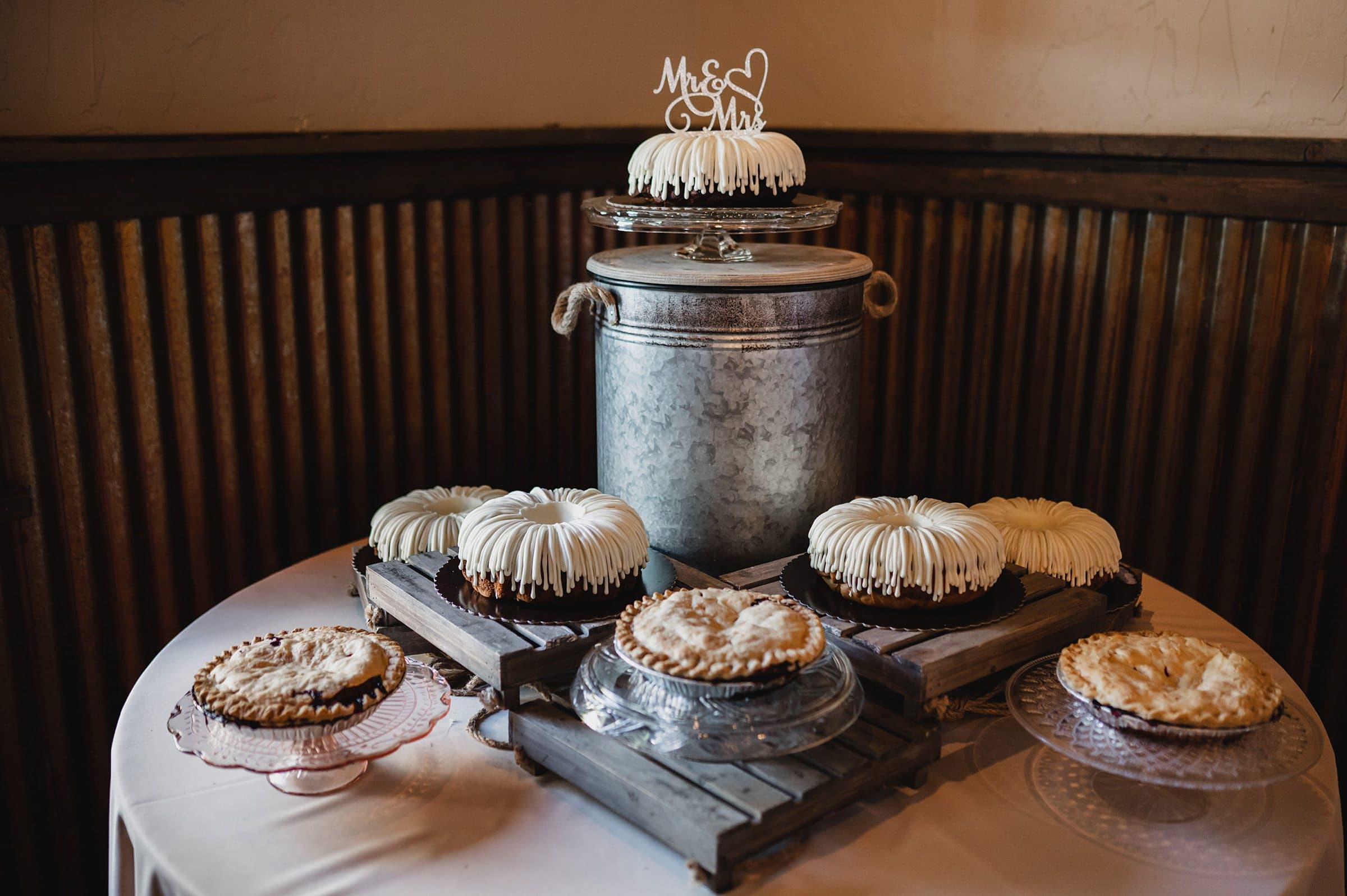 A selection of assorted cakes and pies sits on a table, adorned with a Mr & Mrs topper on one cake, set against the rustic backdrop of a Gorge Crest Vineyard wedding.