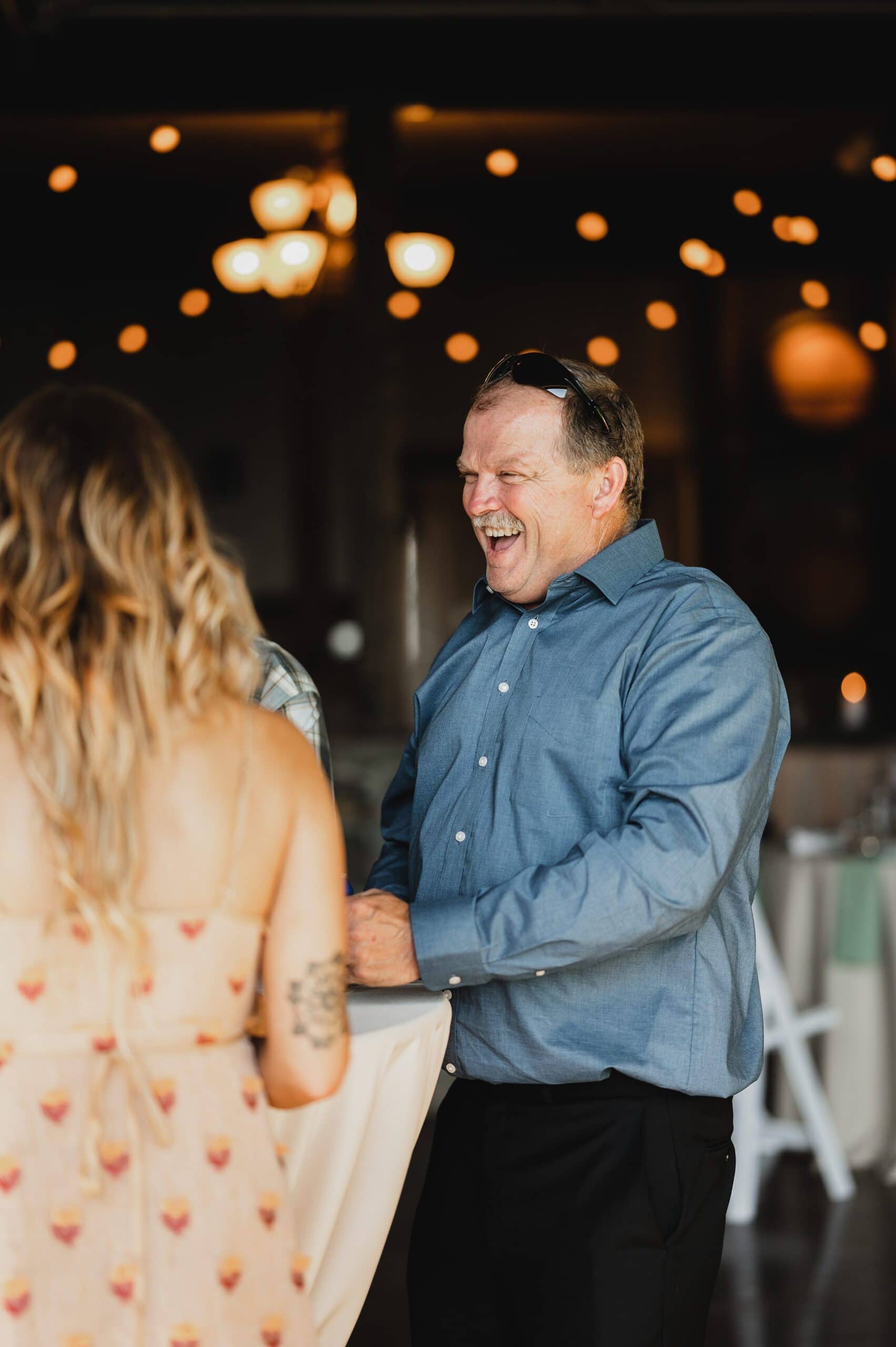 A man in a blue shirt laughs with a woman at a Gorge Crest Vineyard wedding, warm lighting and string lights creating an inviting backdrop.