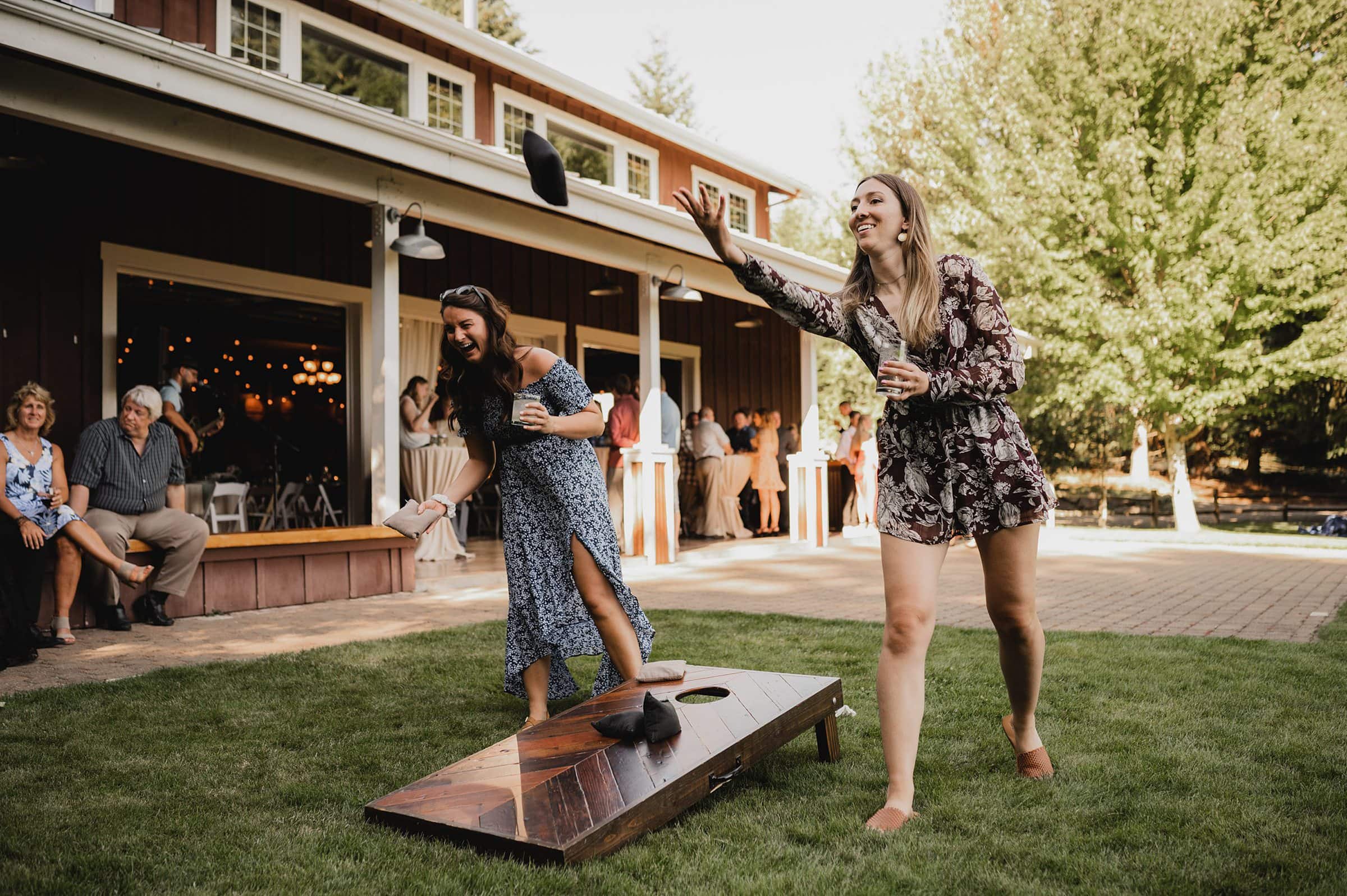 At a gathering at Gorge Crest Vineyards, two women play cornhole outdoors as onlookers sit nearby, capturing a joyful moment reminiscent of a lively wedding celebration.