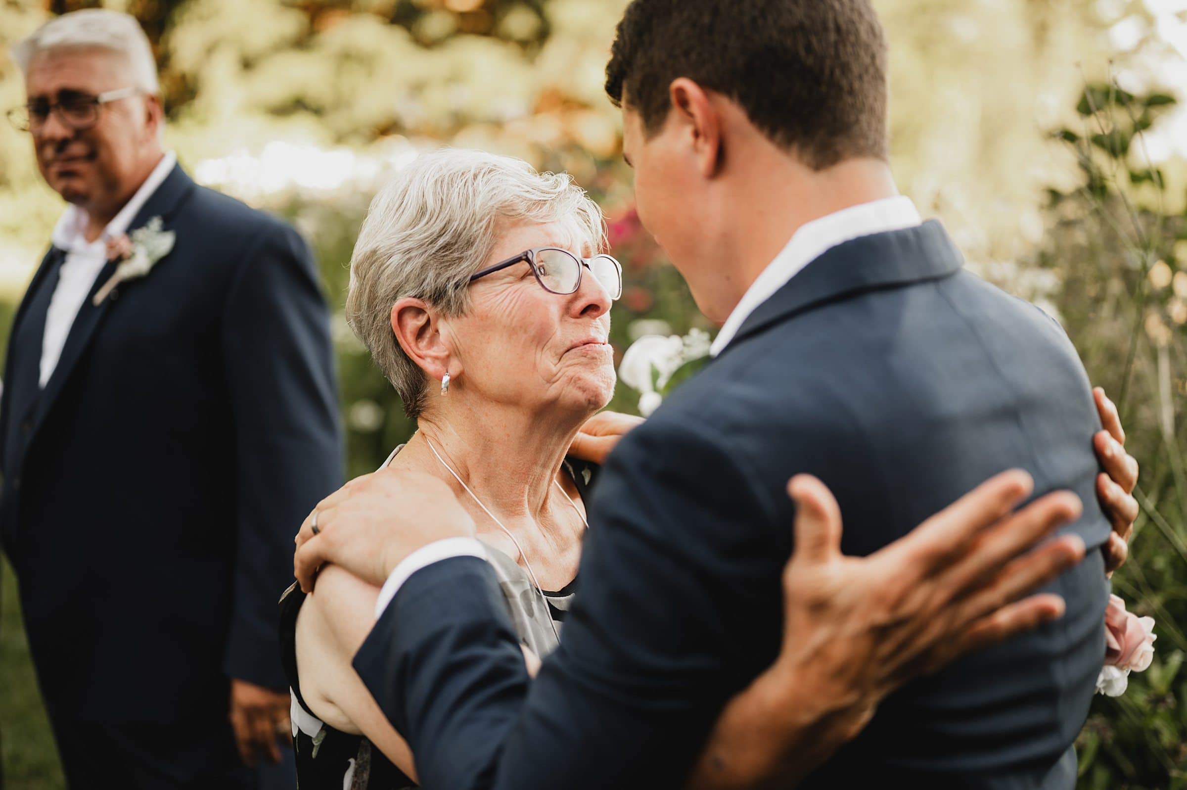 An older woman and a younger man share a tender moment outdoors at Gorge Crest Vineyard, capturing a heartfelt scene while another man stands in the background.