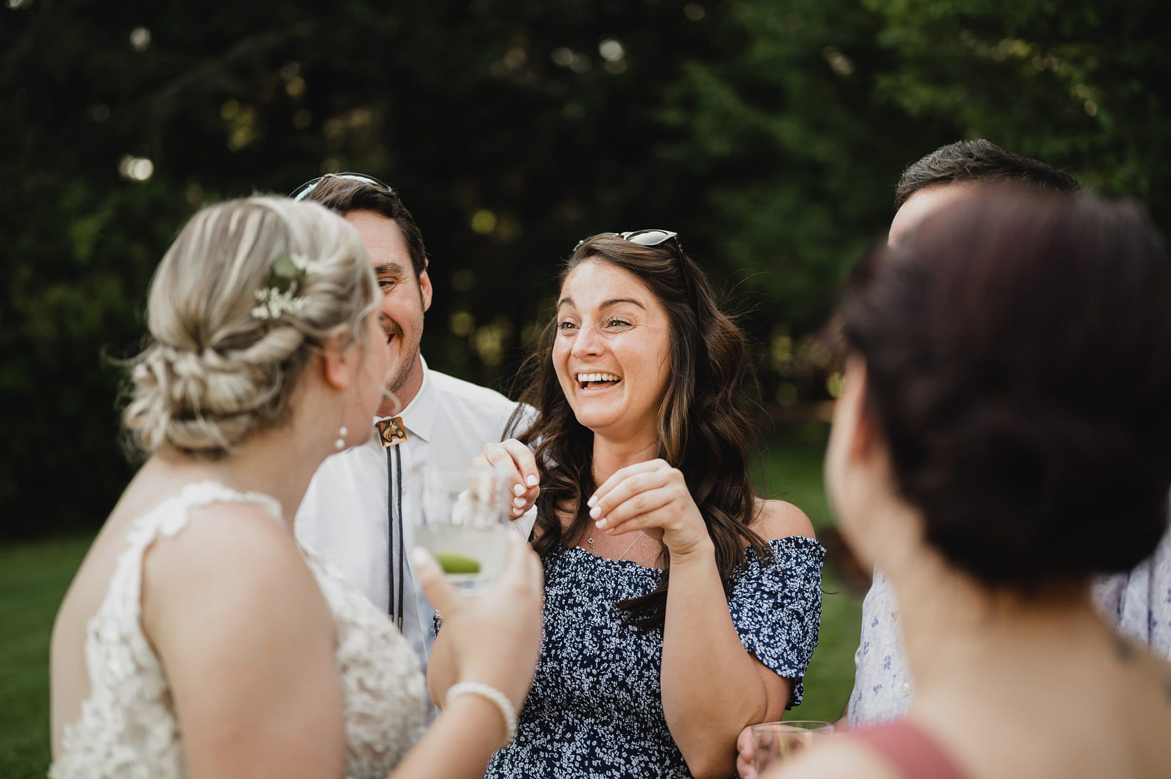 People smiling and talking outdoors, one woman holding a drink, with trees in the background at a picturesque Gorge Crest Vineyard wedding.