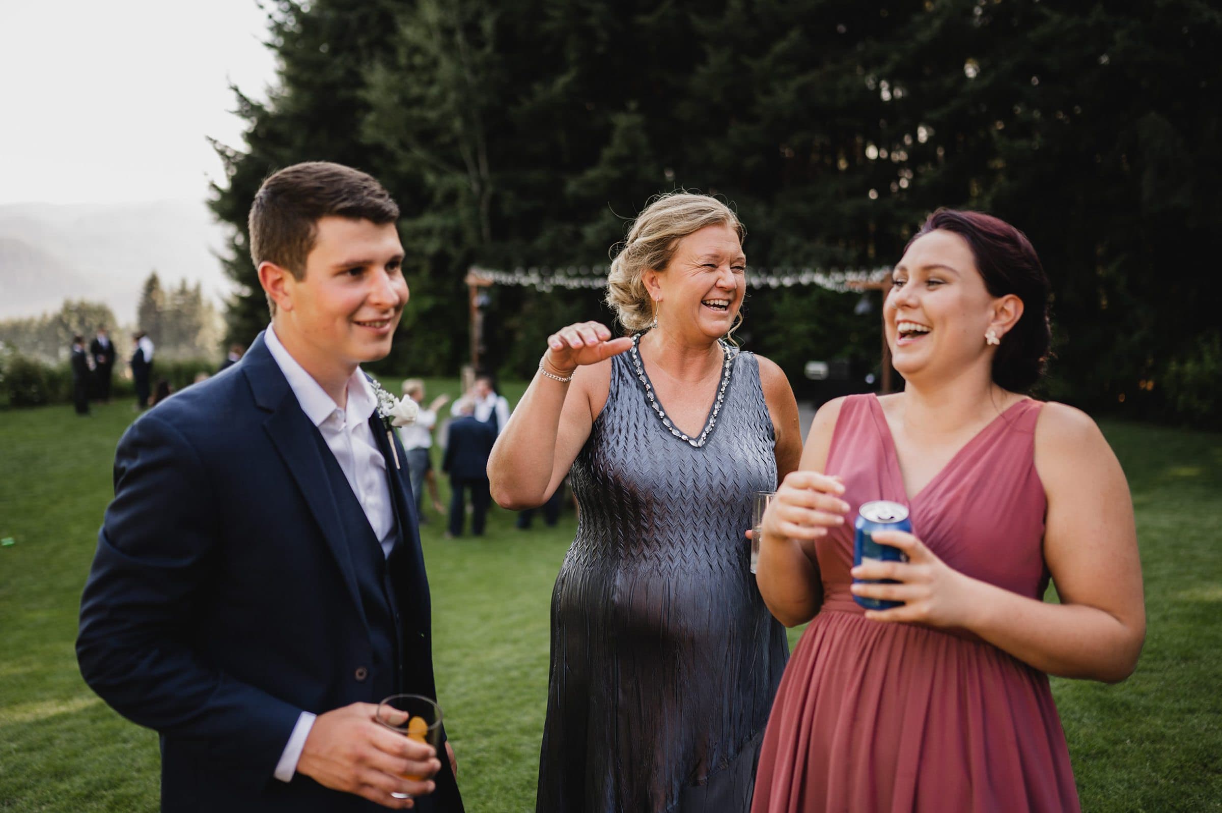 Three people laughing outdoors at a formal event, holding drinks amidst the beautiful trees in the background, capture the joy of a Gorge Crest Vineyards wedding.