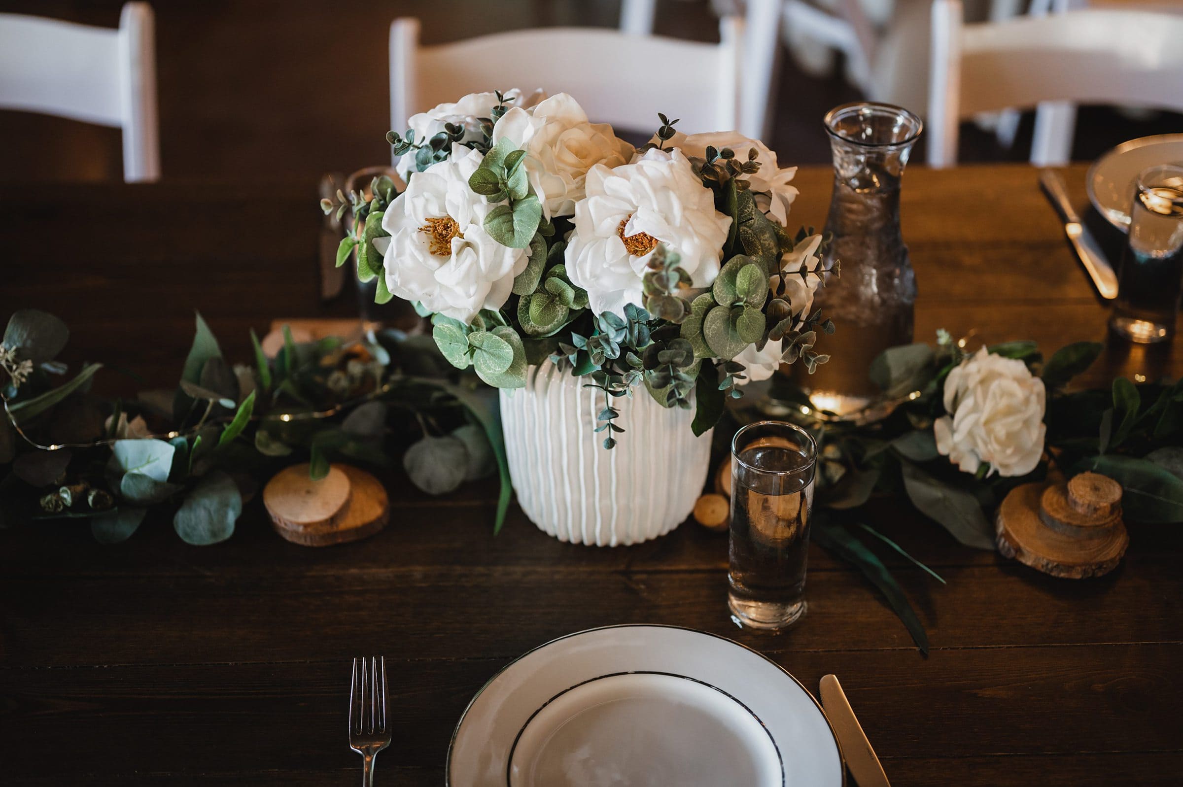 White flowers and greenery in a ceramic vase sit on a wooden table, accompanied by a garland and elegant table setting. Perfect for capturing stunning wedding photos at Gorge Crest Vineyards.