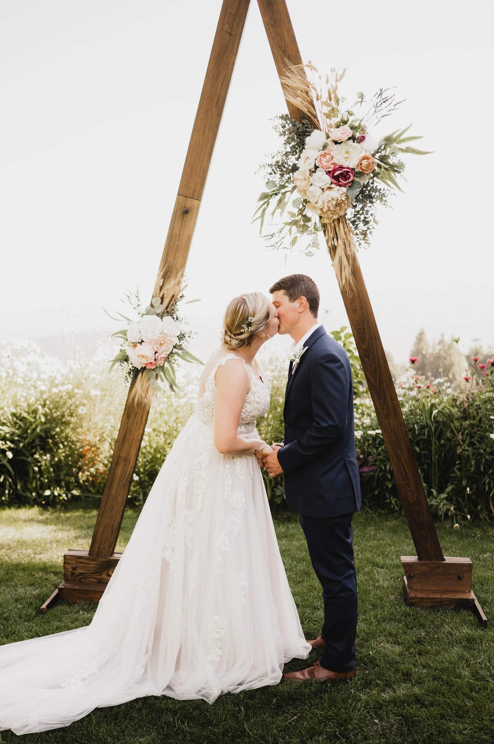 A bride and groom kiss in front of a wooden triangle arch adorned with flowers outdoors, capturing a beautiful moment for their wedding photos at Gorge Crest Vineyards.