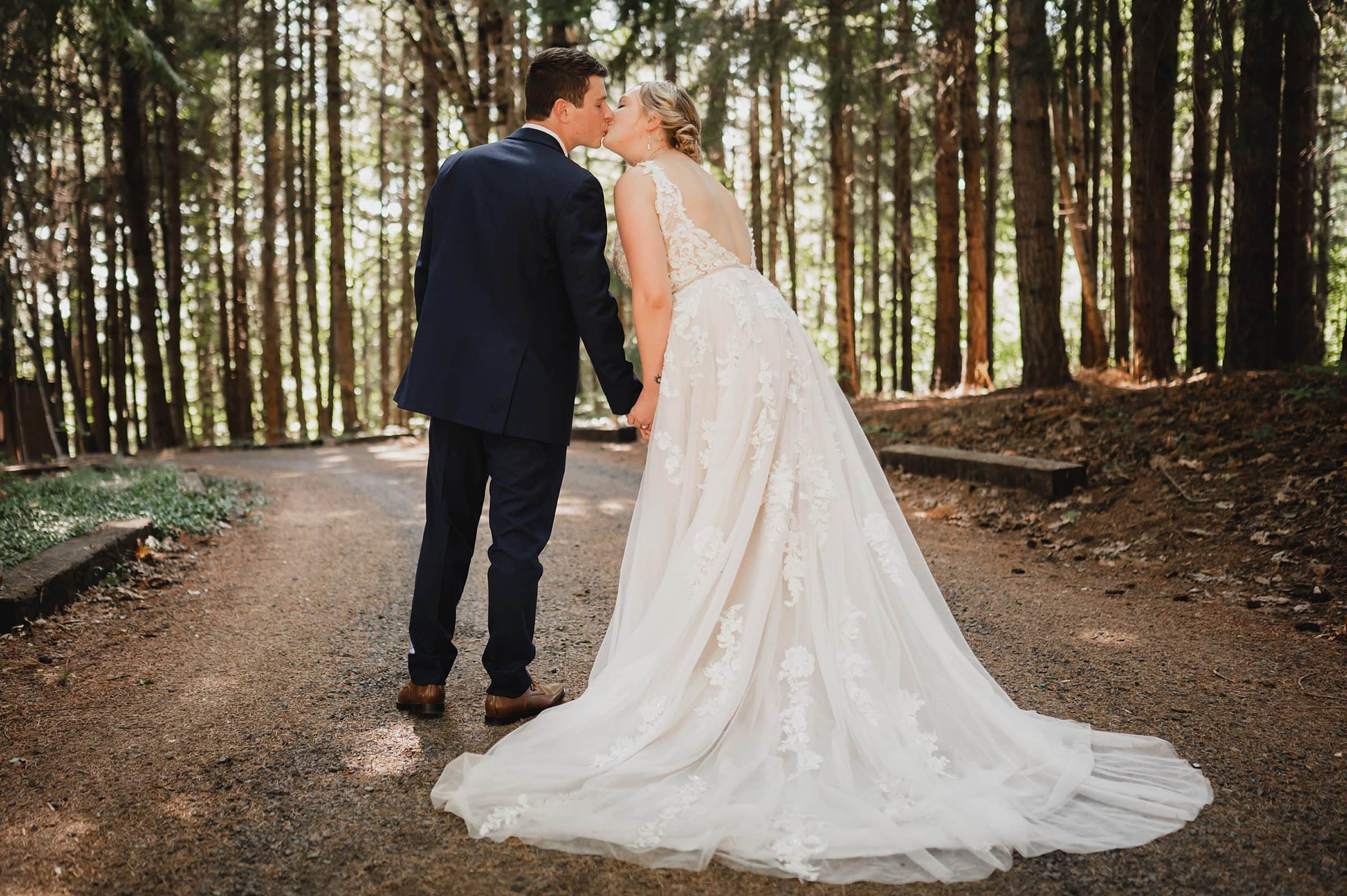 Amidst the enchanting forest path, the bride and groom share a kiss, their hands intertwined, with trees lining the background. This magical moment captures the romance akin to a Gorge Crest Vineyards wedding, where natures beauty sets a captivating scene.