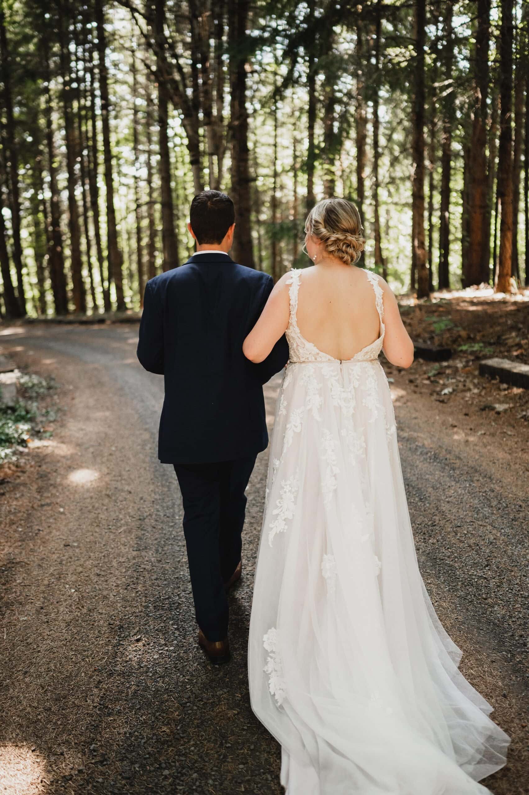 The bride and groom walk hand in hand down a forest path at Gorge Crest Vineyards, with sunlight filtering through the trees.