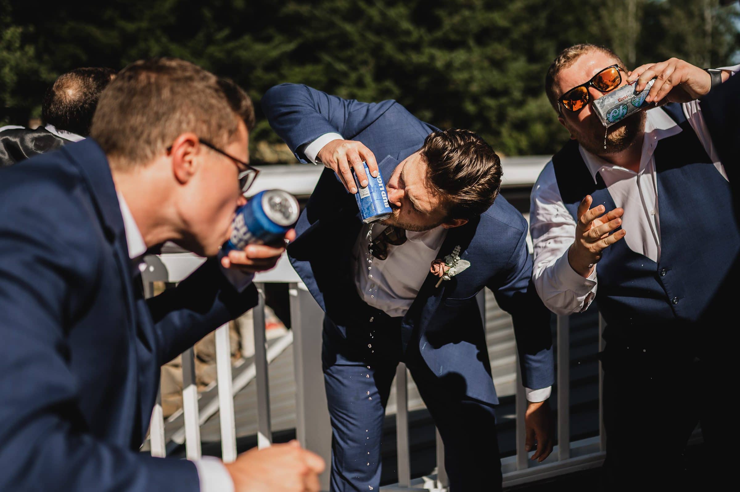 Three men in suits energetically drinking from cans on a sunny day, set against the lush greenery at a Gorge Crest Vineyards wedding.