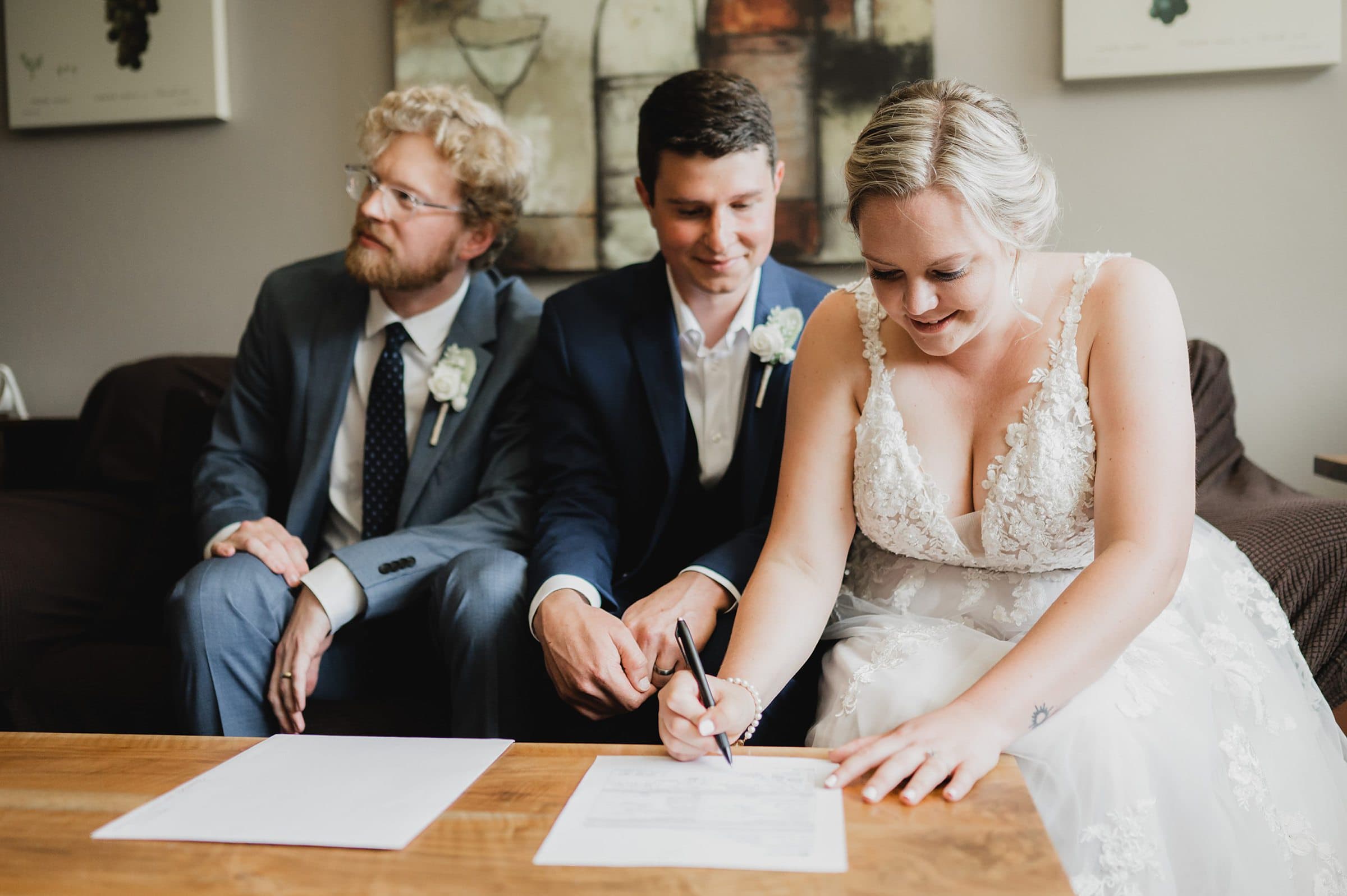 At Gorge Crest Vineyard, a bride in a white dress signs a document at a table, while two men in suits sit beside her, capturing the perfect wedding moment.