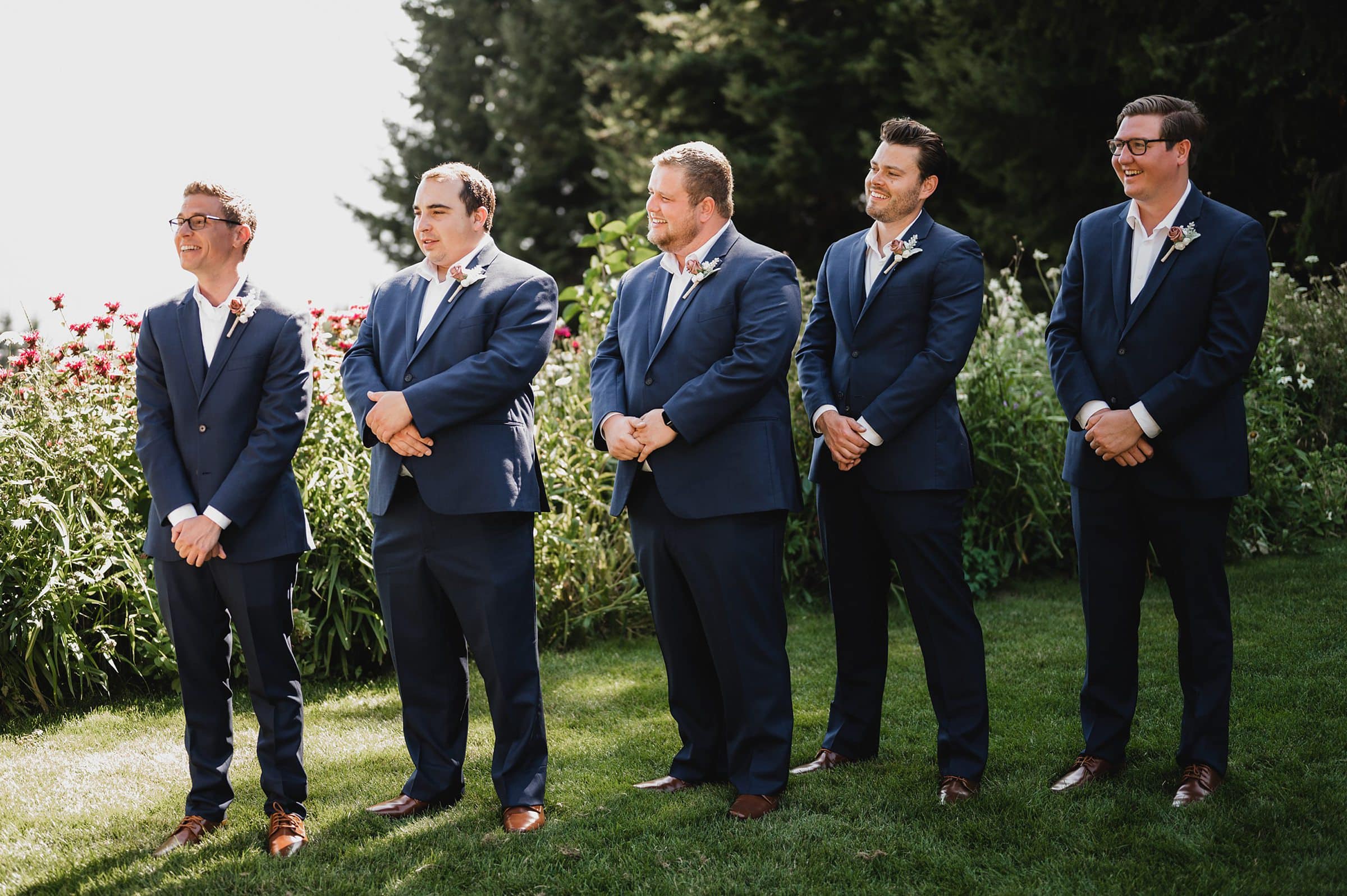 Five groomsmen in blue suits stand on grass near flowering bushes, smiling under the daylight at Gorge Crest Vineyards wedding.