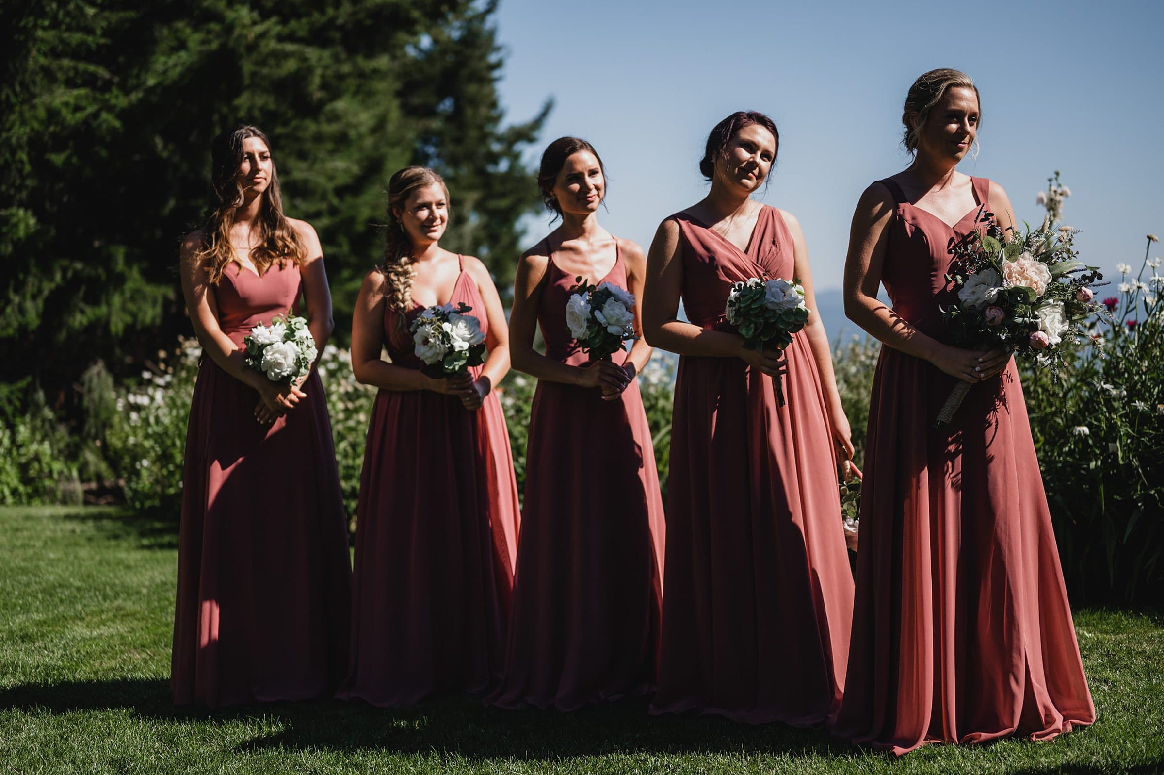 Five women in matching pink dresses stand outdoors, holding bouquets against a backdrop of greenery. This picturesque scene beautifully captures moments from a Gorge Crest wedding.